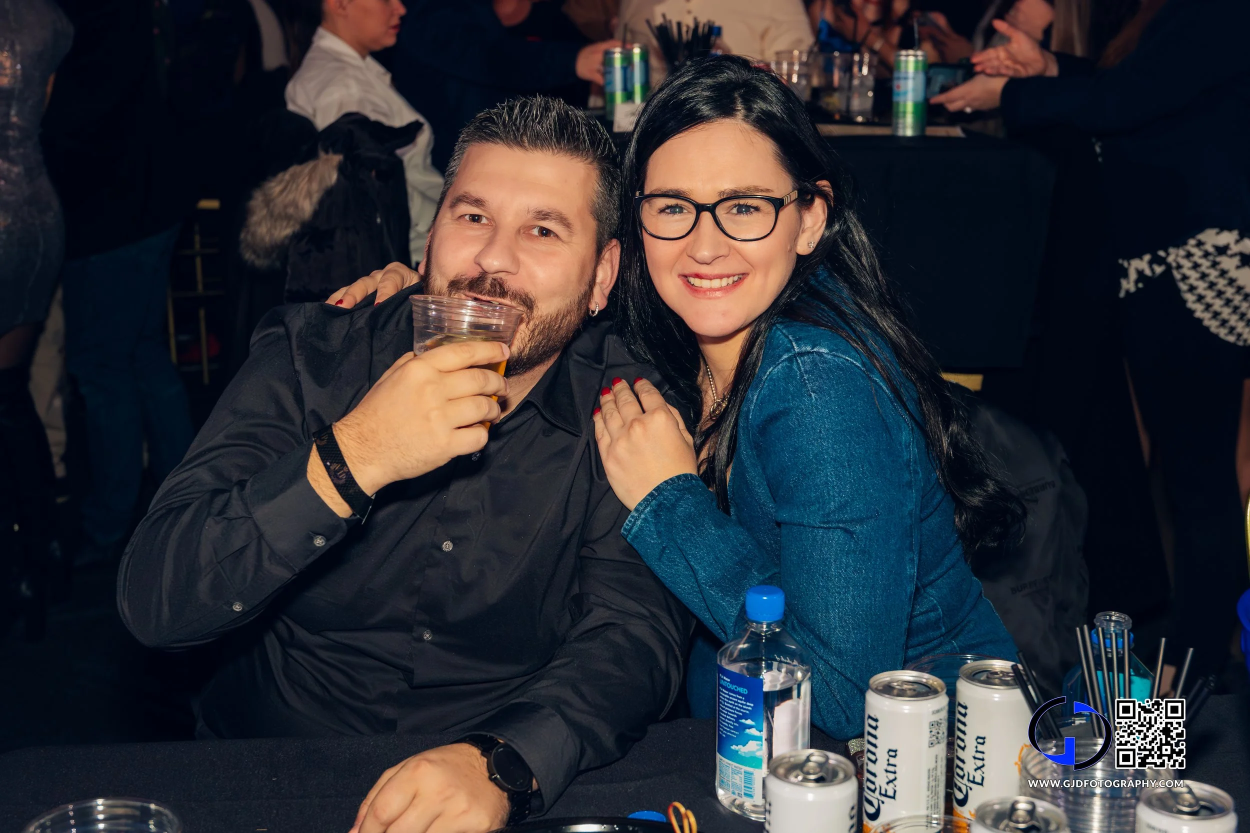 A man and woman sitting at a bar or party table, smiling and posing for the camera. The man is holding a drink, and the woman has her hand on his shoulder. There are cans of Corona beer and a water bottle on the table.