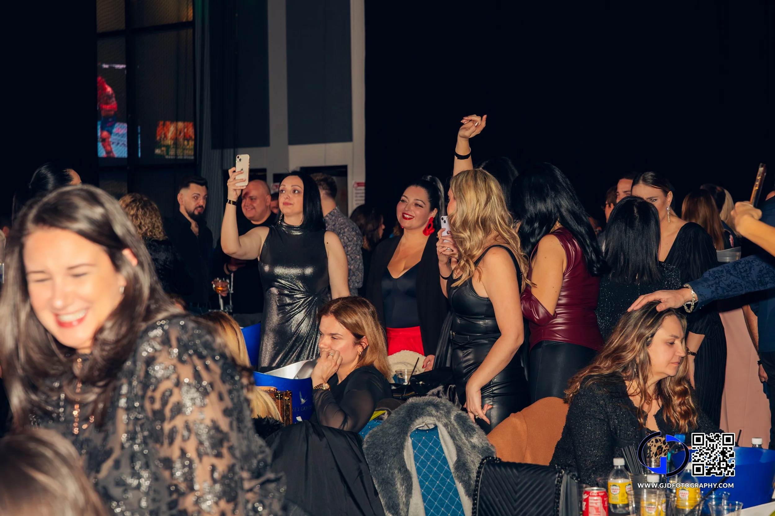Group of women at a formal event, some taking photos with smartphones, others smiling, wearing elegant dresses, in a dimly lit venue.