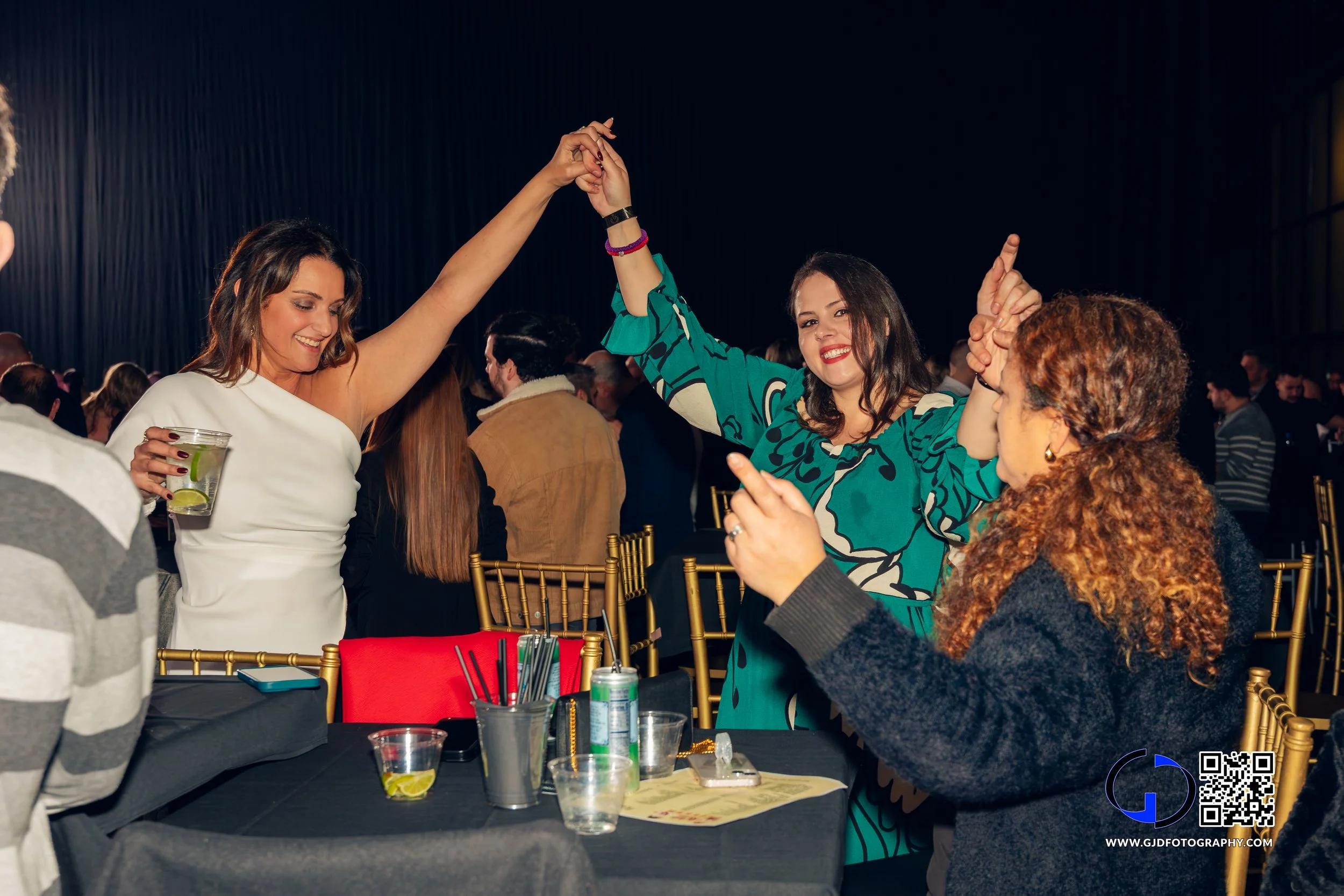 Two women dancing and smiling at a lively social event, with a table of drinks and party supplies in the foreground.