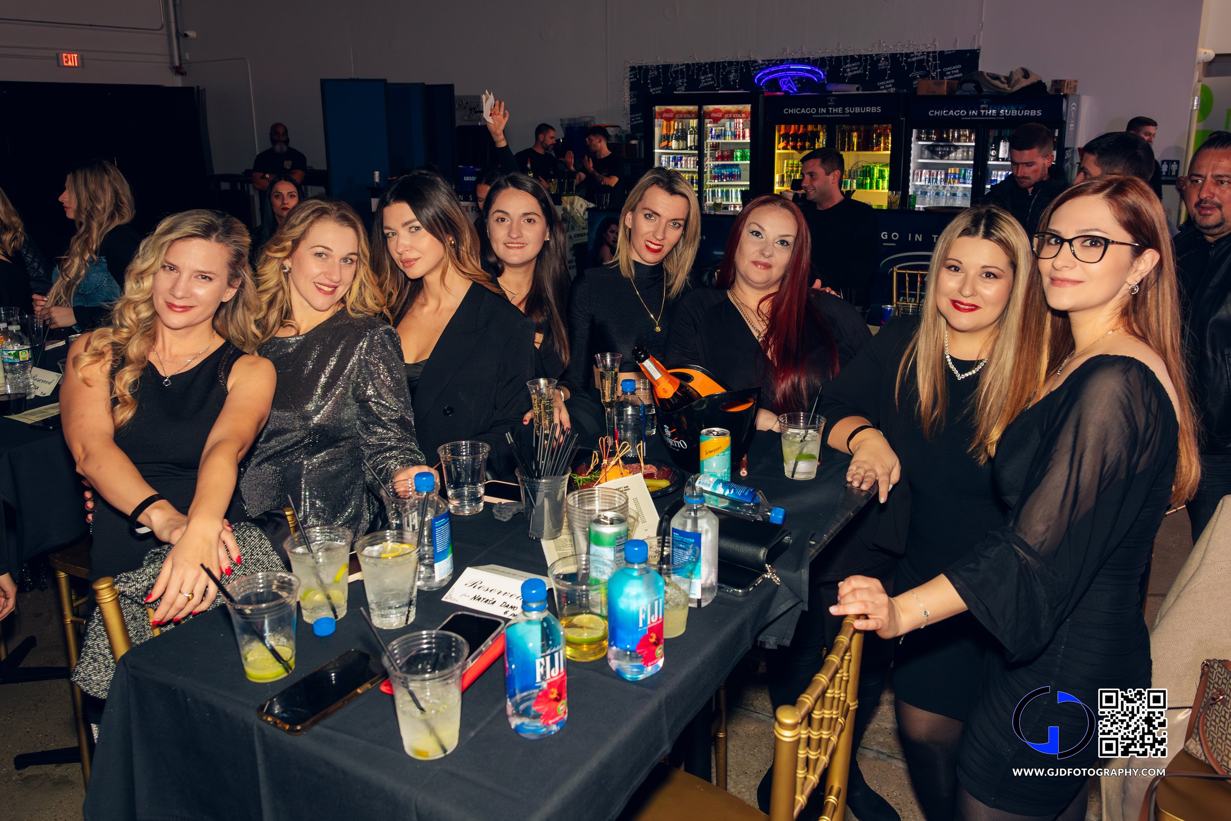 A group of women at a celebratory event around a table with drinks and snacks, smiling at the camera.