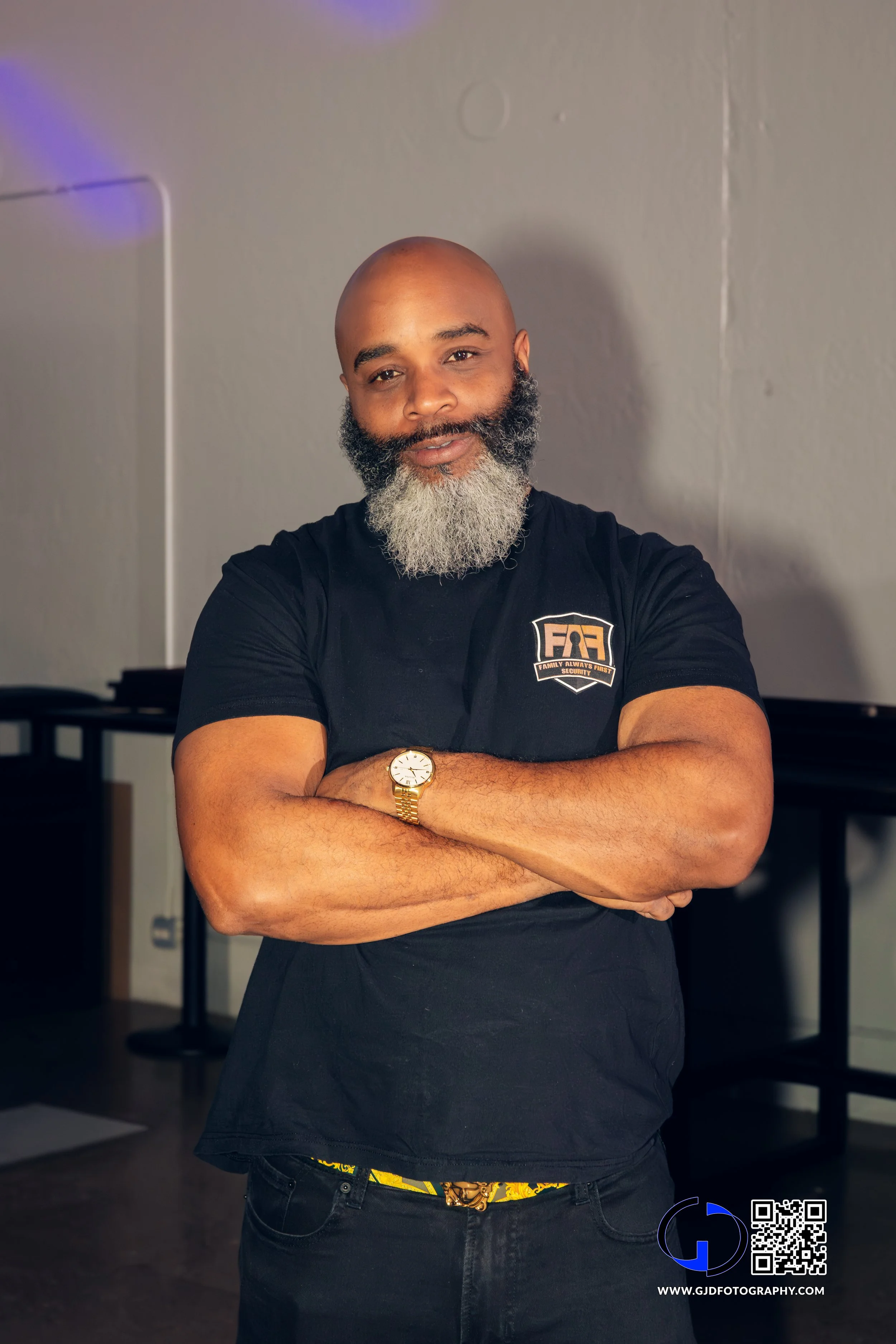 A man with a beard, wearing a black t-shirt with a logo on it, standing with arms crossed inside a room. He is wearing a gold watch and has a yellow patterned belt showing slightly above his waistband.