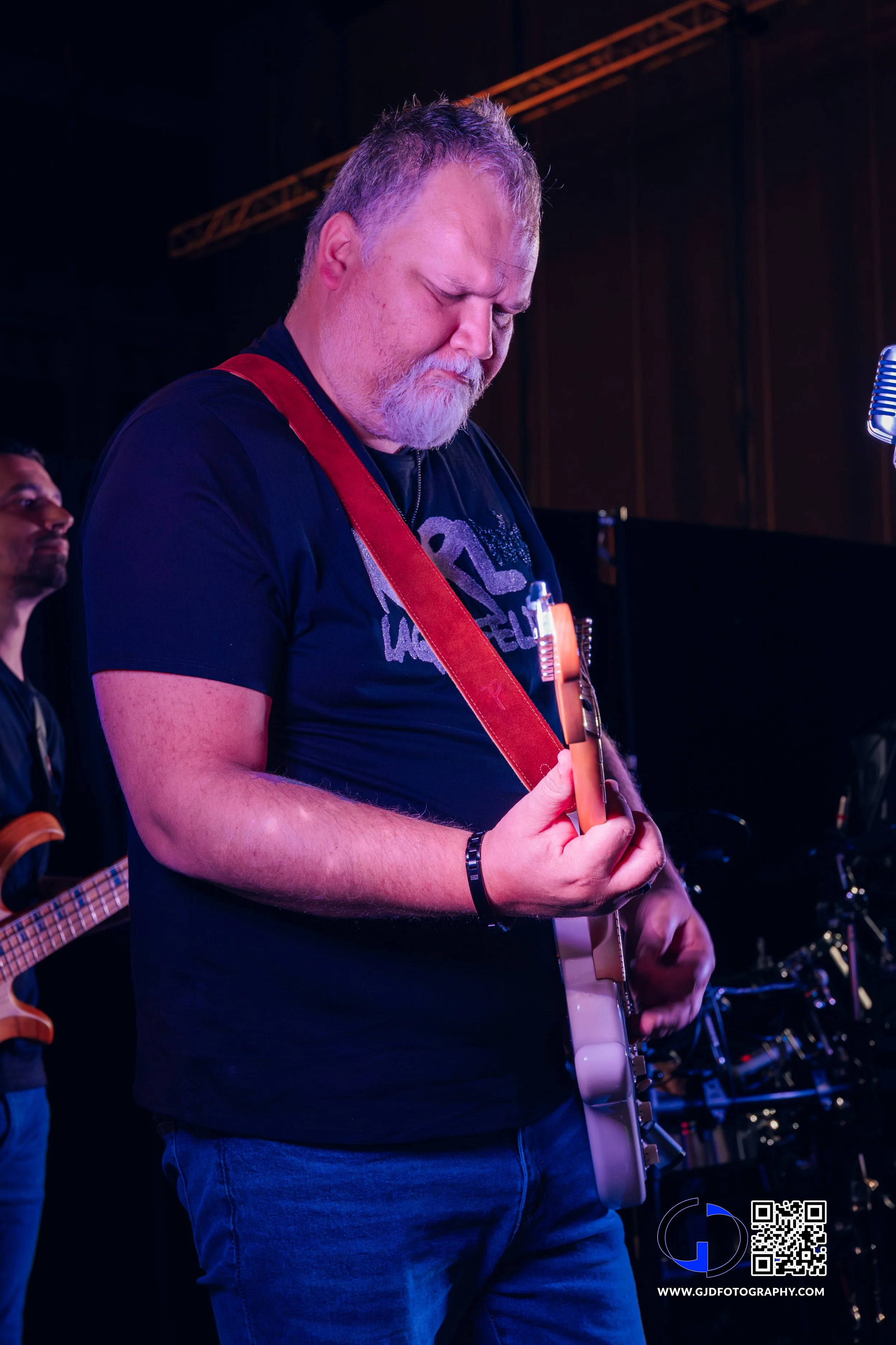 A man with gray hair and beard playing electric guitar during a performance on stage, with another musician in the background and stage equipment around.