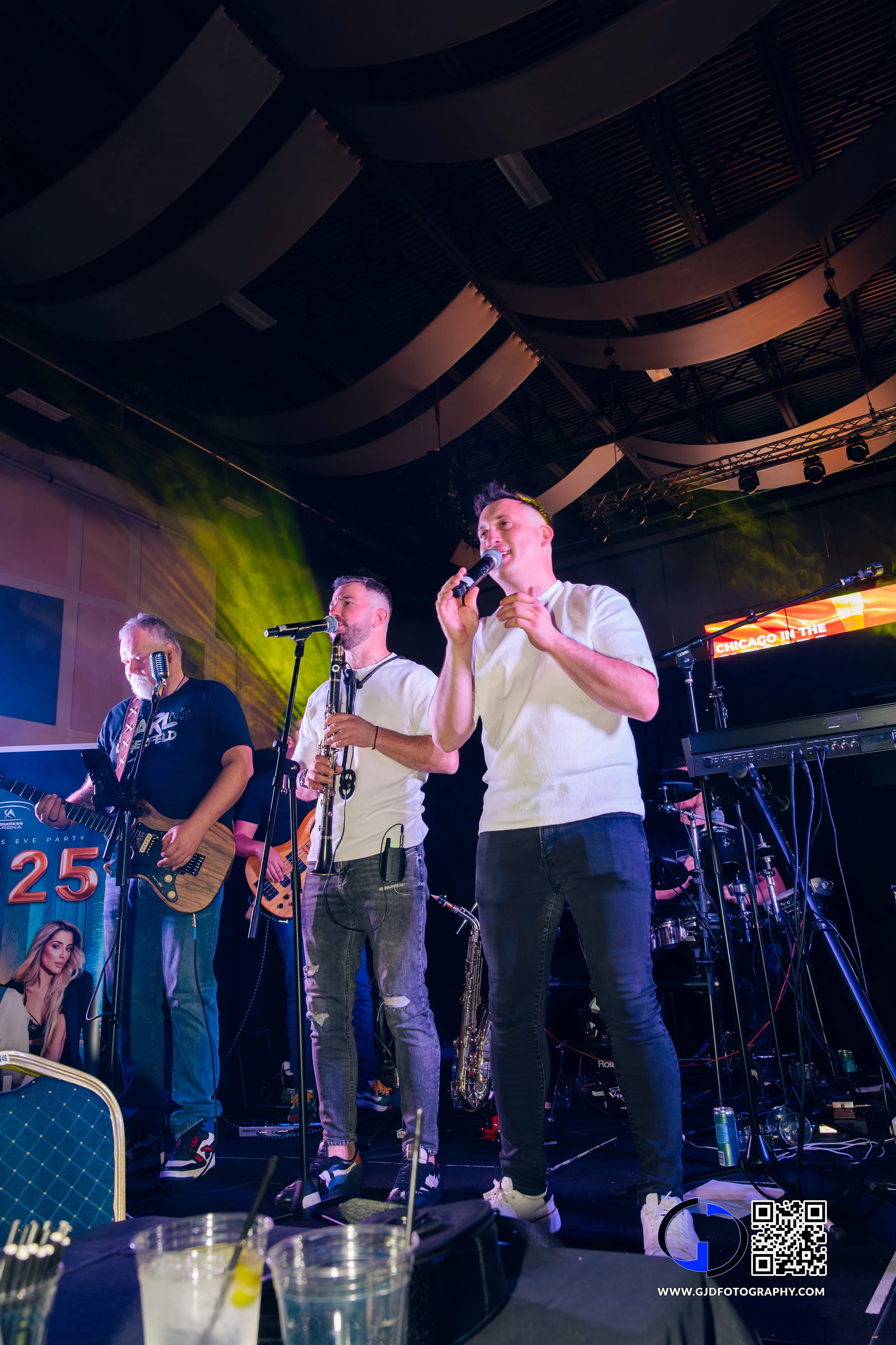 A band of four male musicians performing on stage with microphones, guitars, and a keyboard, under colorful stage lighting at a concert or live event.