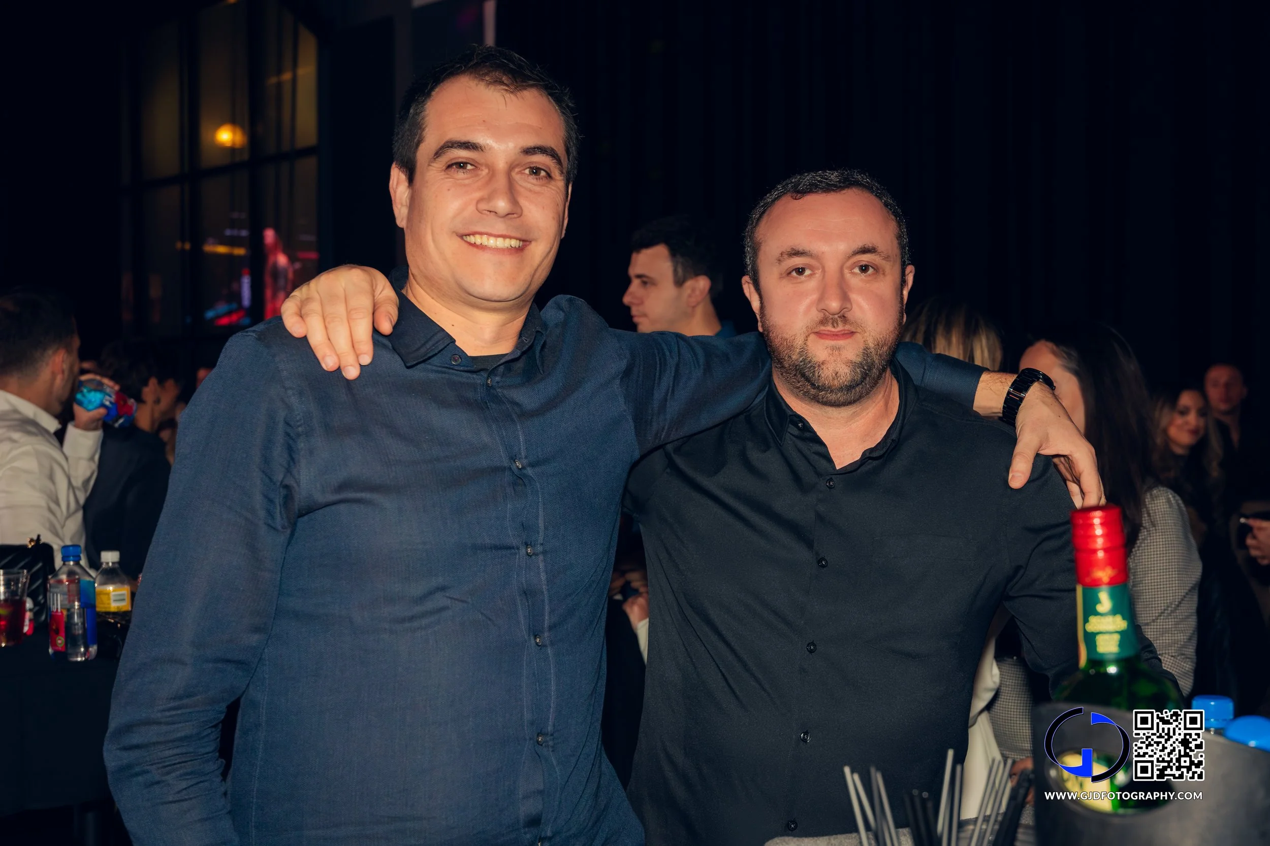 Two men smiling and posing together at a social event, with one arm around each other's shoulders, in a dimly lit venue with other people in the background.