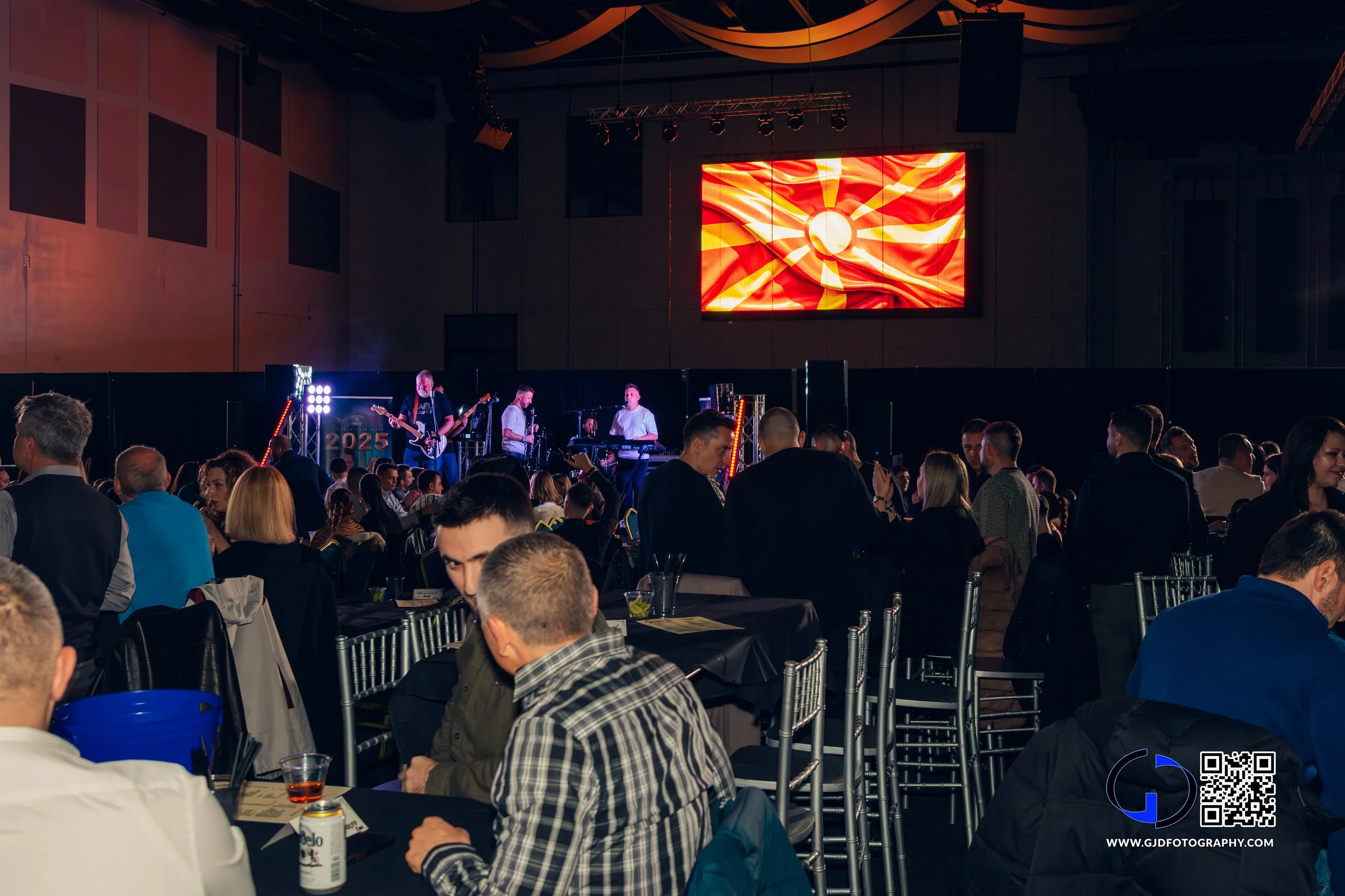 Crowd attending a live music event in a large indoor venue with a stage and band performing, with a digital screen displaying a red and yellow abstract design behind the performers.