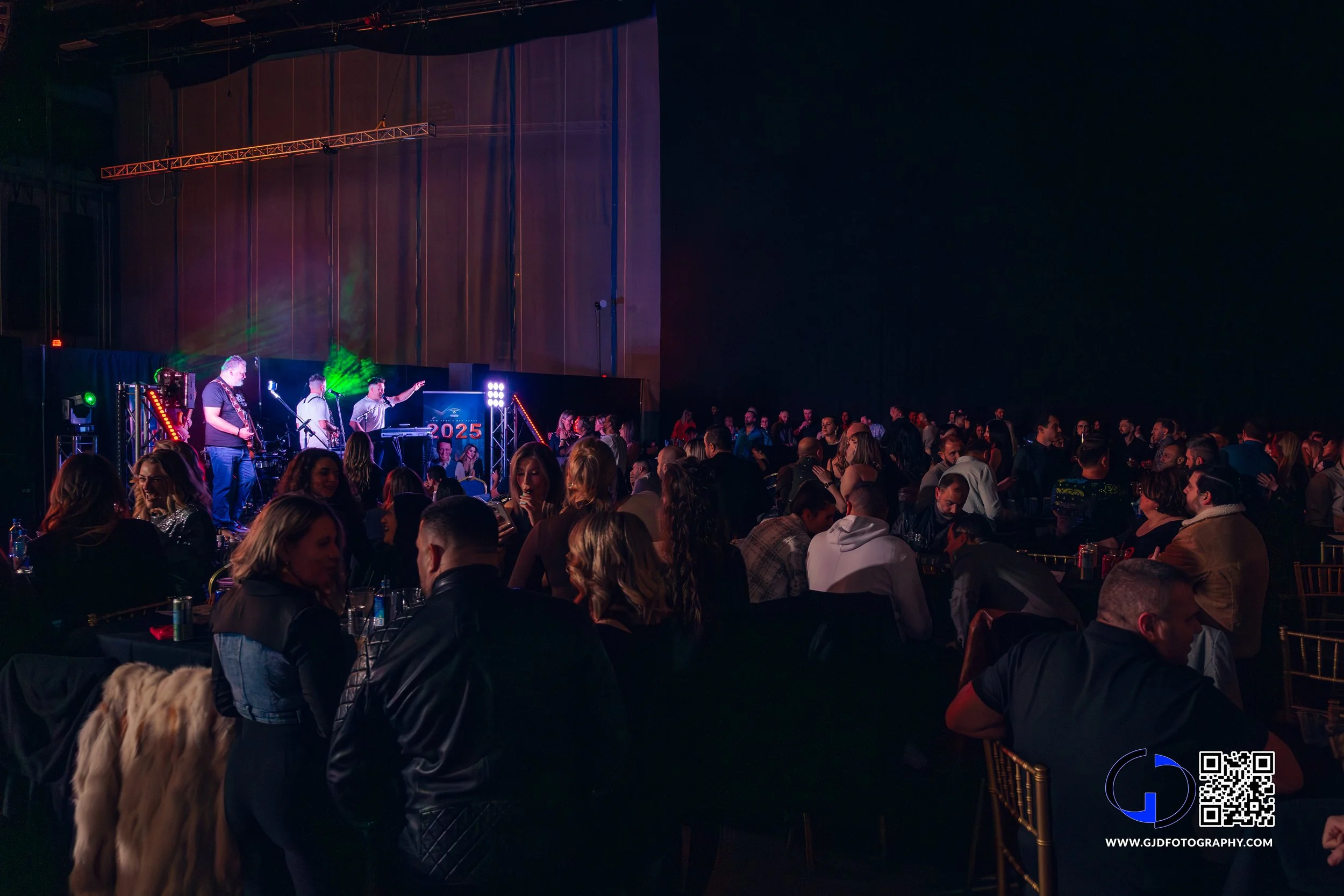 Crowd attending an indoor concert or event with a live band performing on stage, colorful lighting, and people seated at tables watching the performance.