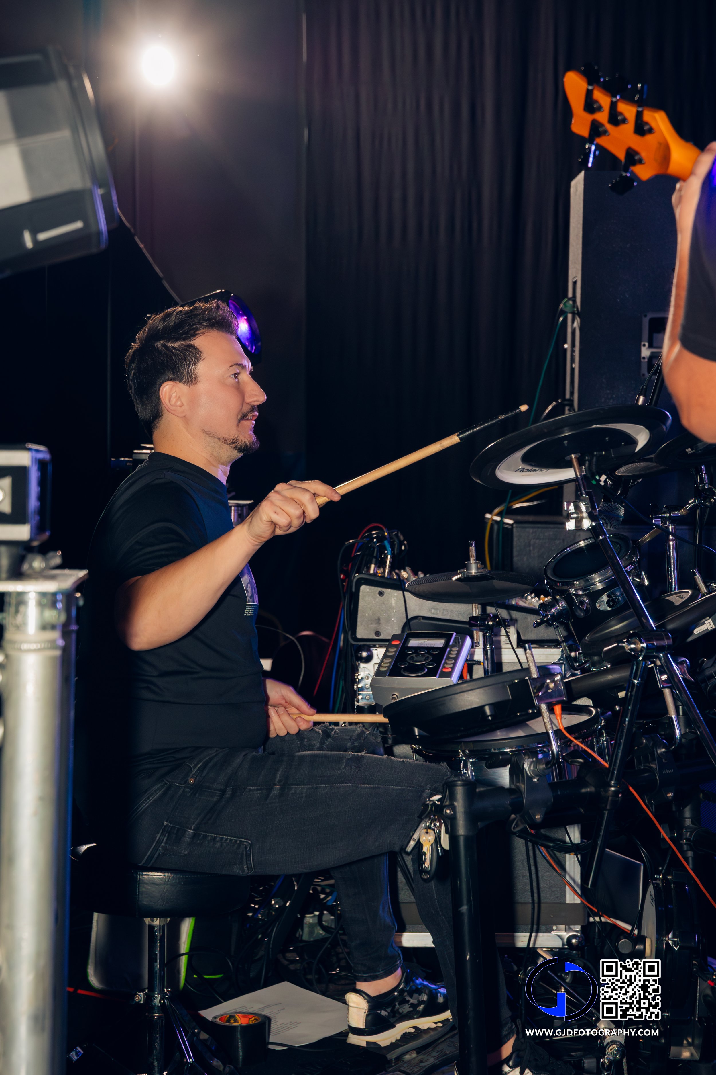 A man playing electronic drums on stage, holding drumsticks with a focused expression, surrounded by musical equipment and a black curtain background.