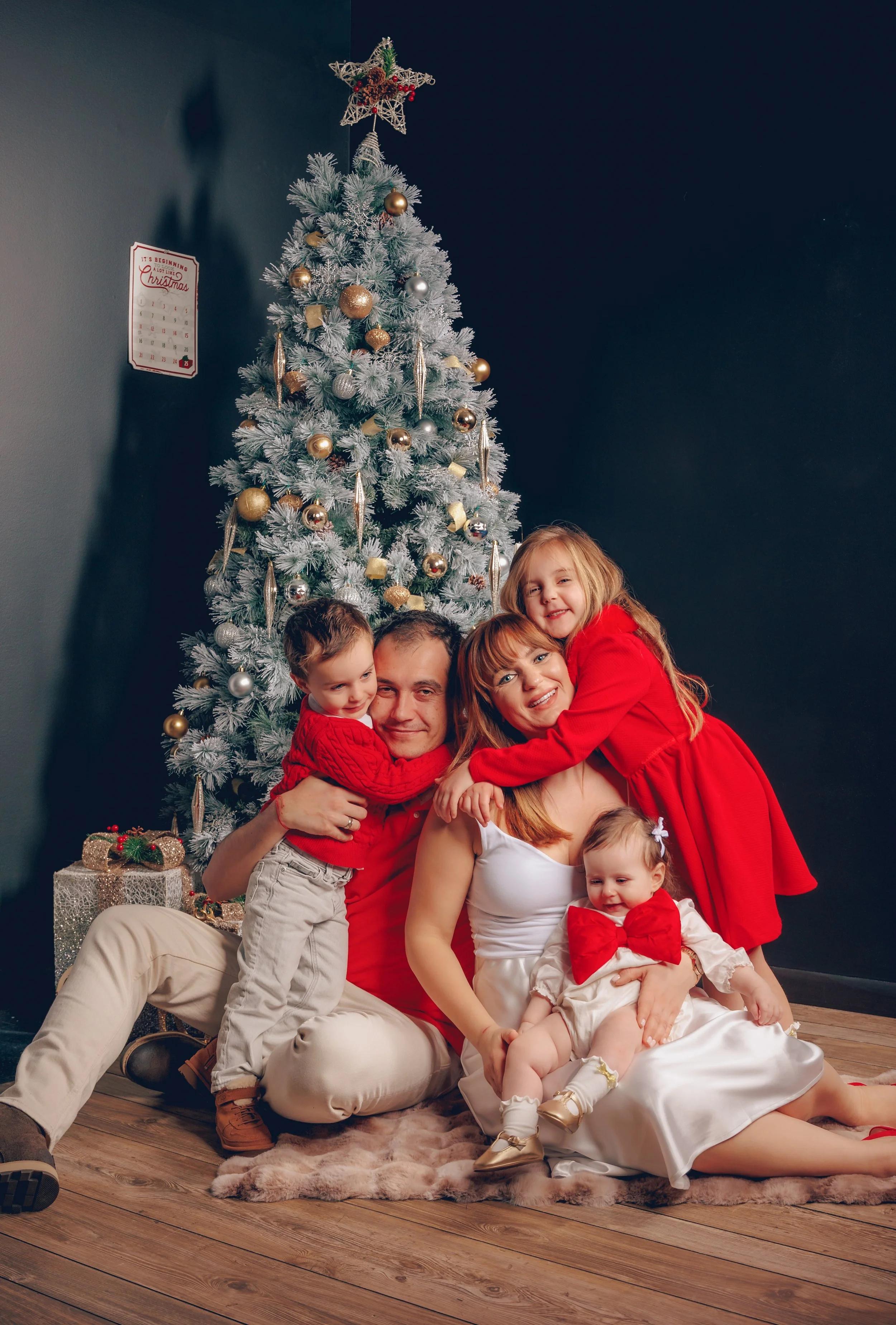 A family of four celebrating Christmas next to a decorated Christmas tree with gifts, all wearing red and white, smiling and hugging each other.