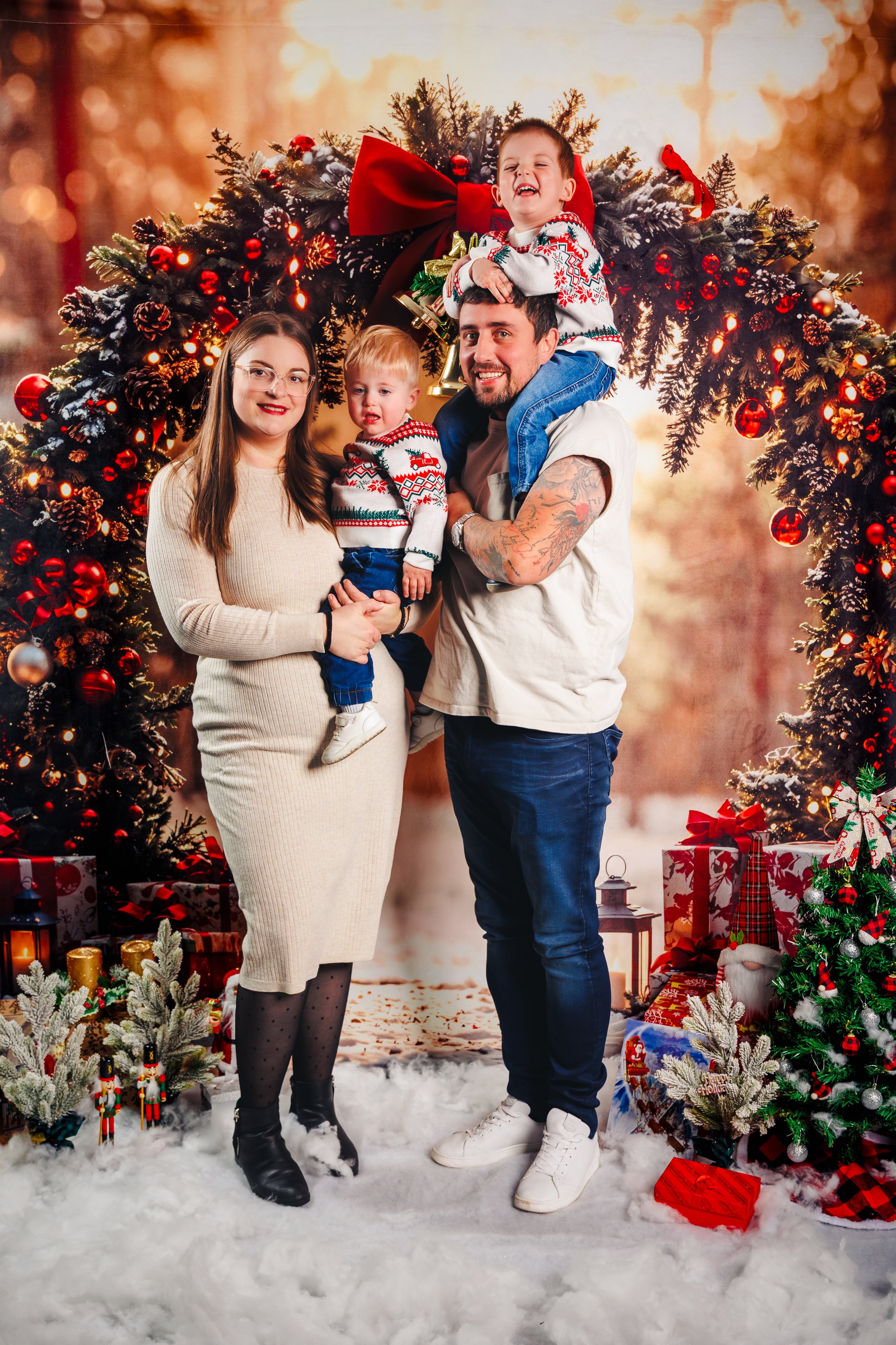 A family of four celebrating Christmas outdoors with decorated trees and wrapped presents, standing on artificial snow.