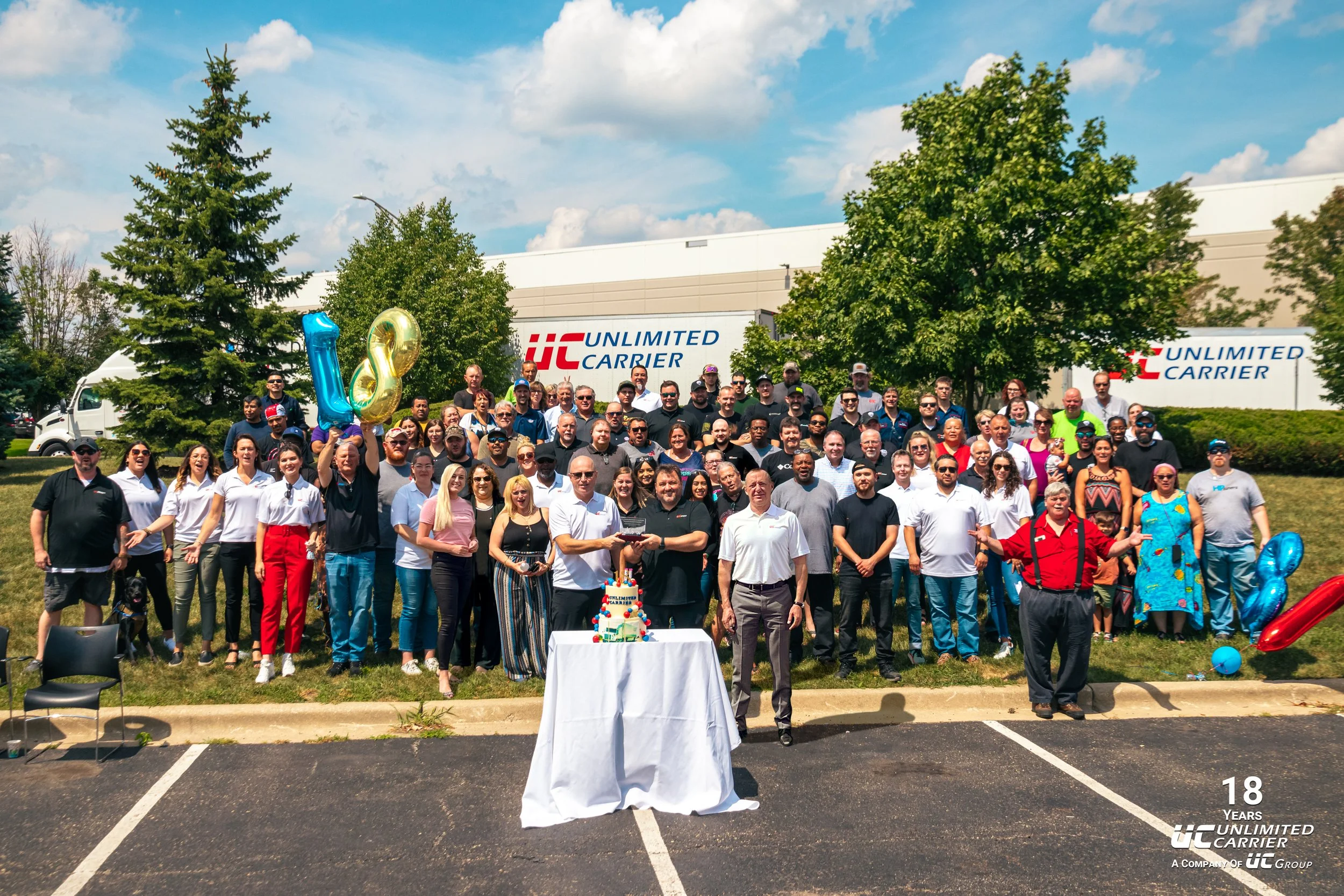 Group photo of employees celebrating 18 years at Unlimited Carrier, with balloons and a cake, outdoors in front of the company building.