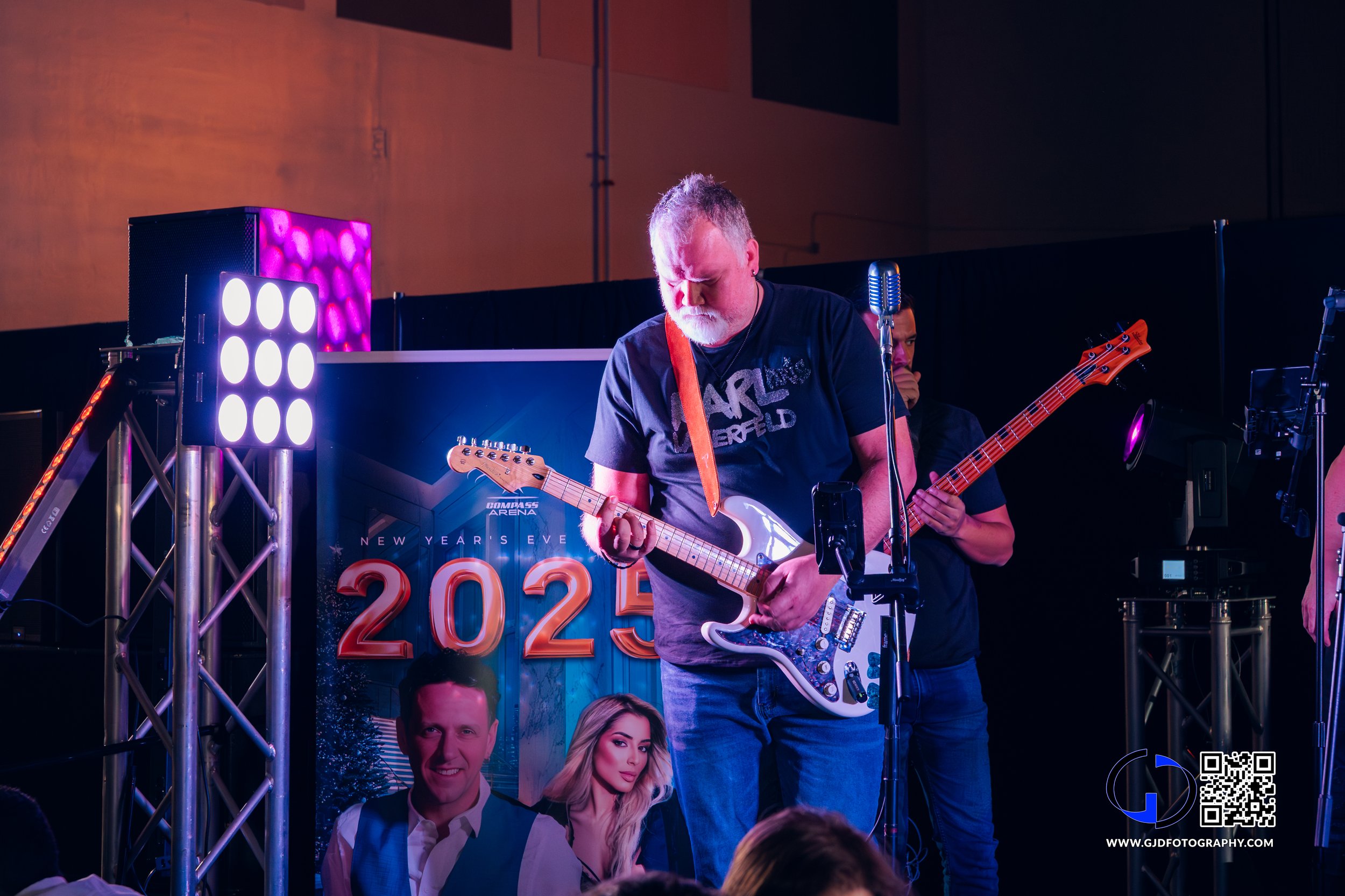 Musician with gray hair and beard playing an electric guitar on stage during a New Year's Eve event in 2023, with stage lights and a poster in the background.