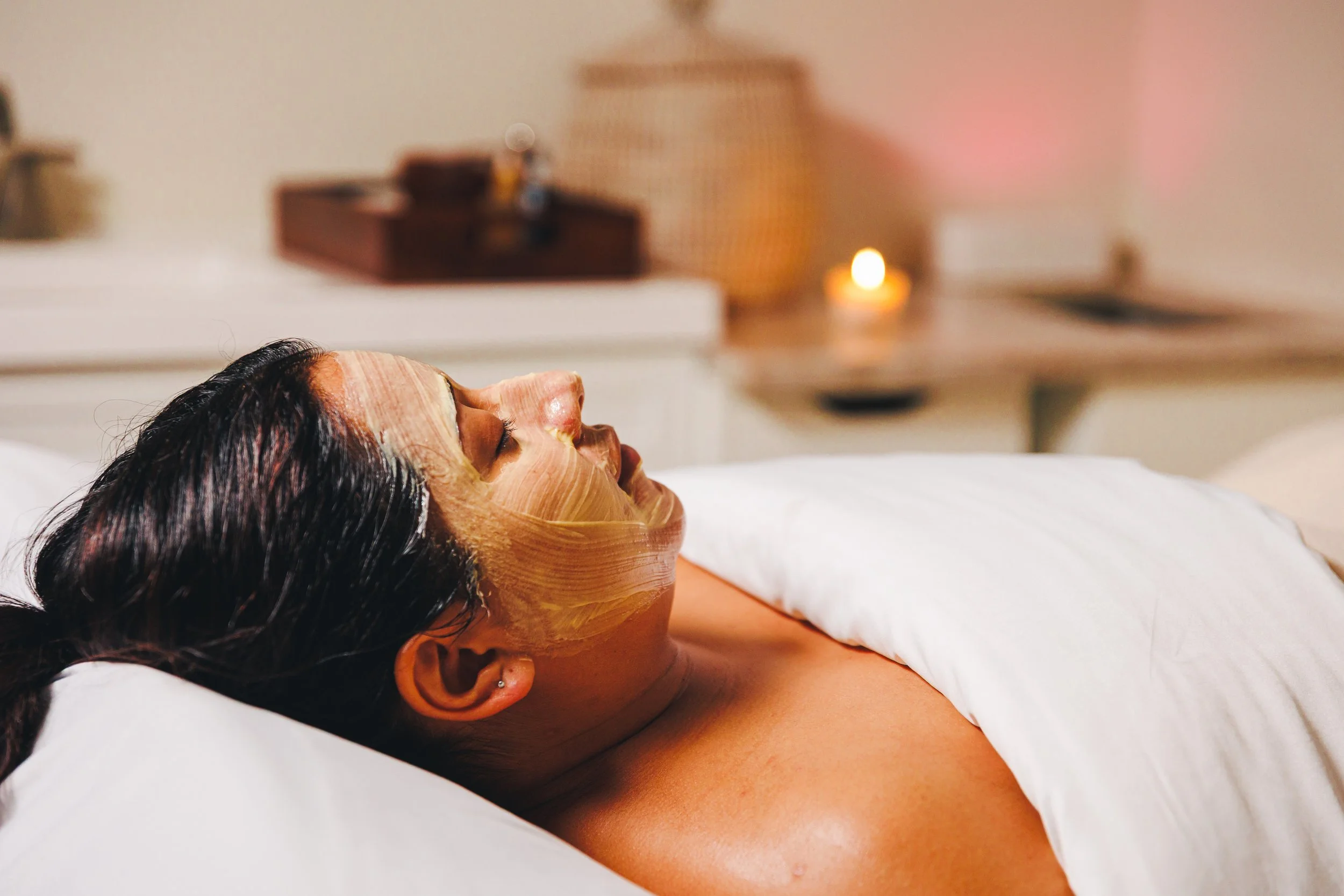 Woman with a facial mask relaxing on a bed with a candle in the background.