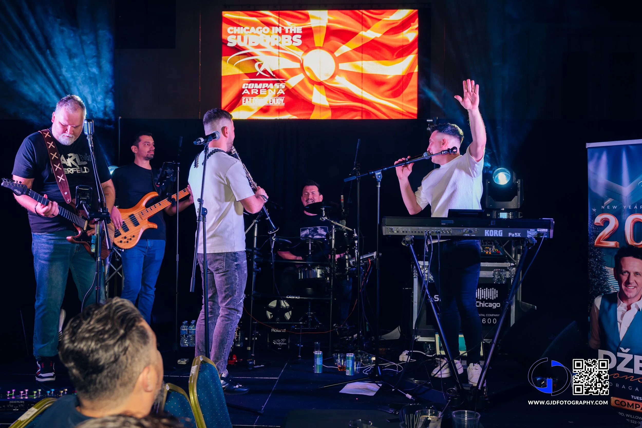 A live band performing on stage with five members, including guitarists, a bassist, a drummer, and a singer, with a large screen above displaying text "Chicago in the Suburbs," at an event celebrating New Year 2020.