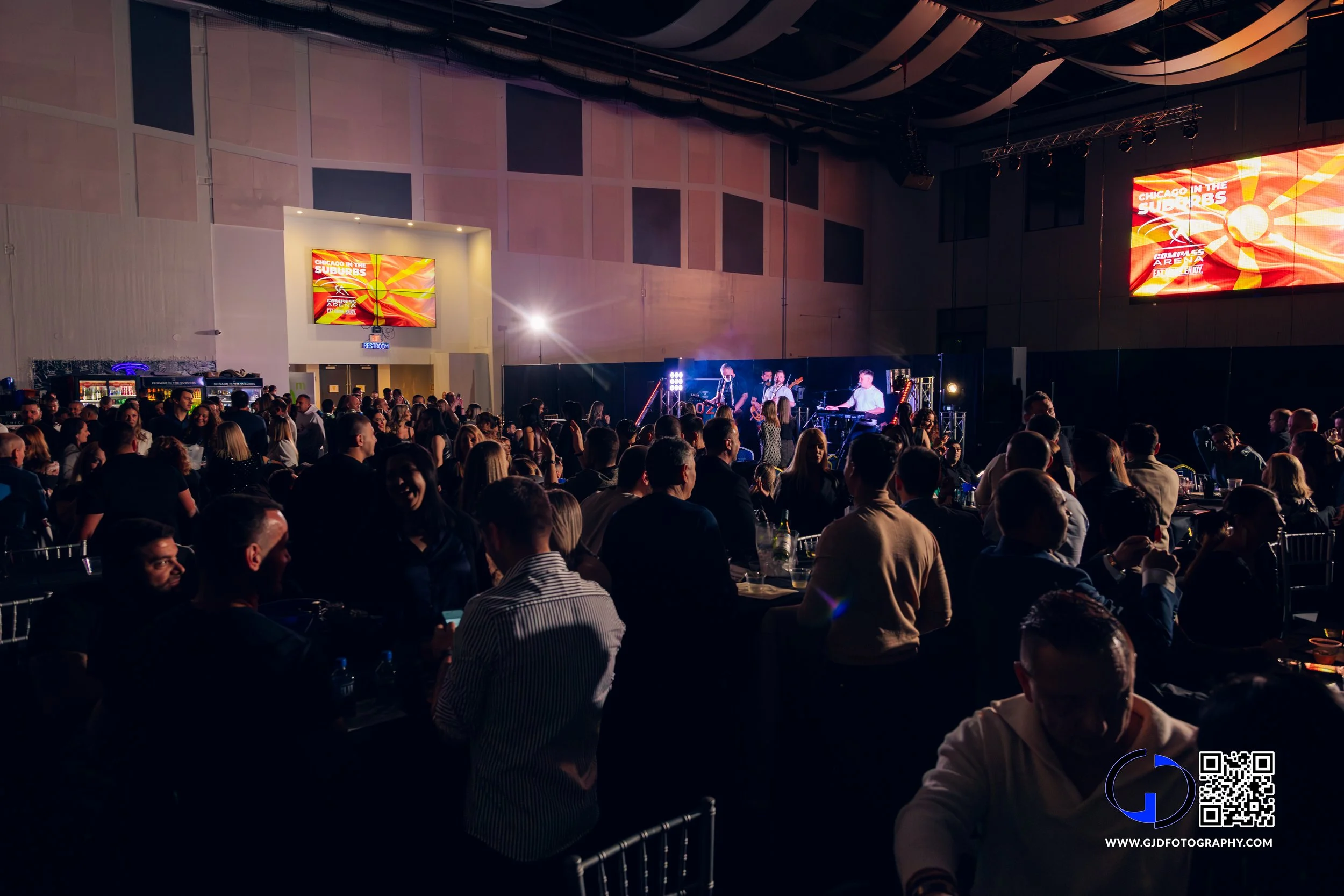Large indoor concert event with a stage, band performing, and audience seated at tables in front.
