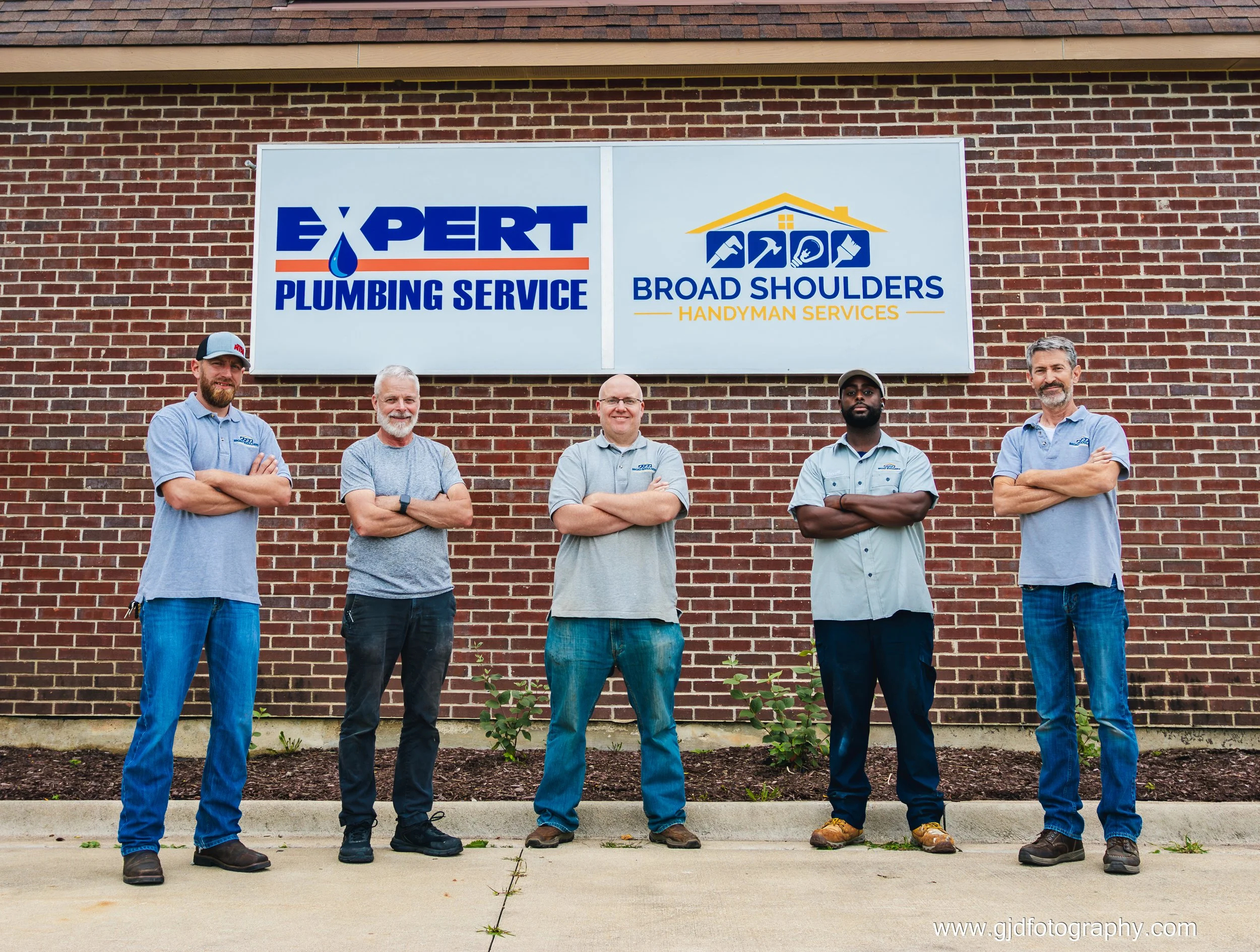 Five men standing with arms crossed in front of a building with two business signs. The left sign reads 'Expert Plumbing Service' with a water droplet logo, and the right sign reads 'Broad Shoulders Handyman Services' with house and tools icons. The 