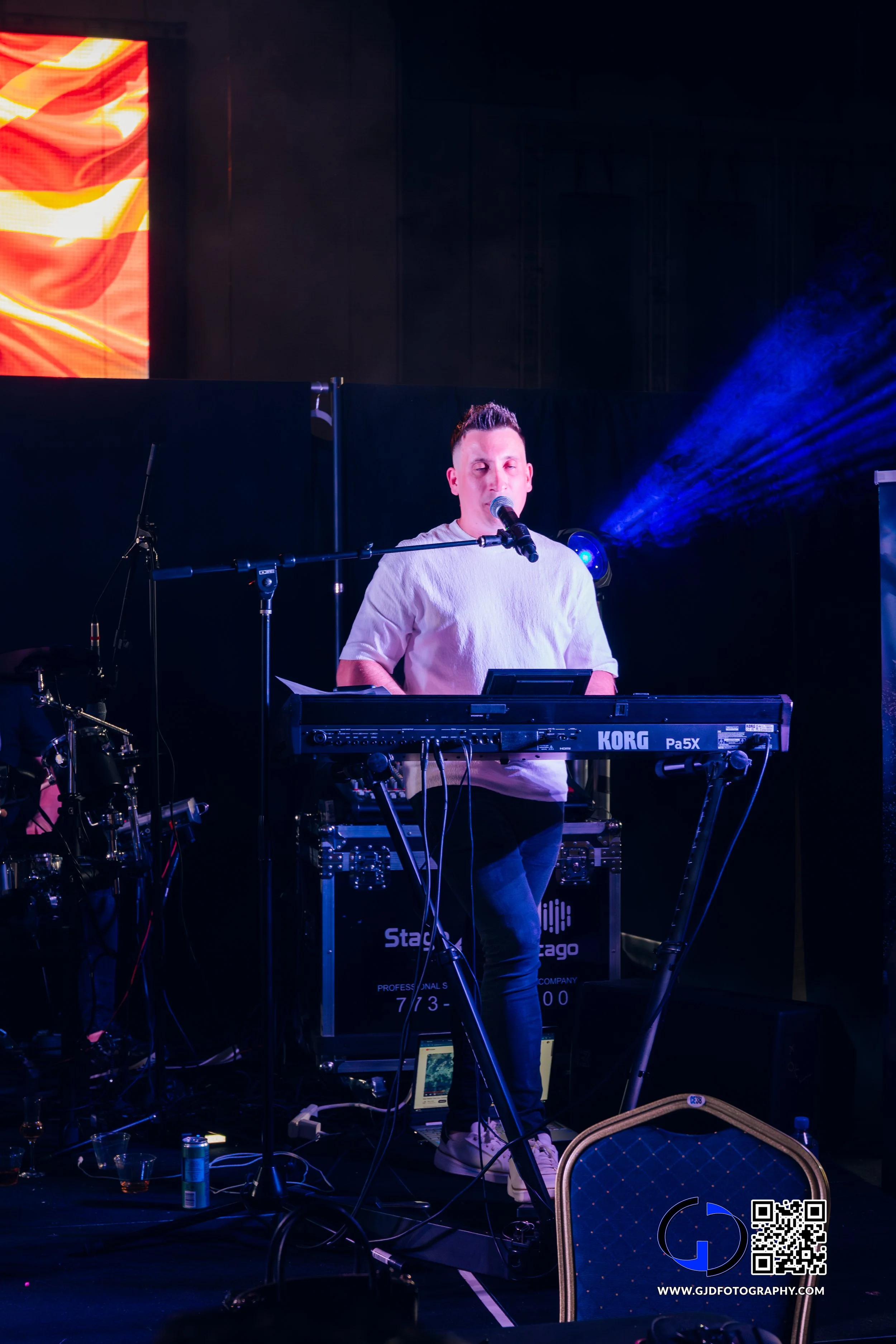 A musician playing a keyboard and singing into a microphone on stage during a live performance, with blue stage lighting and a large LED screen in the background.