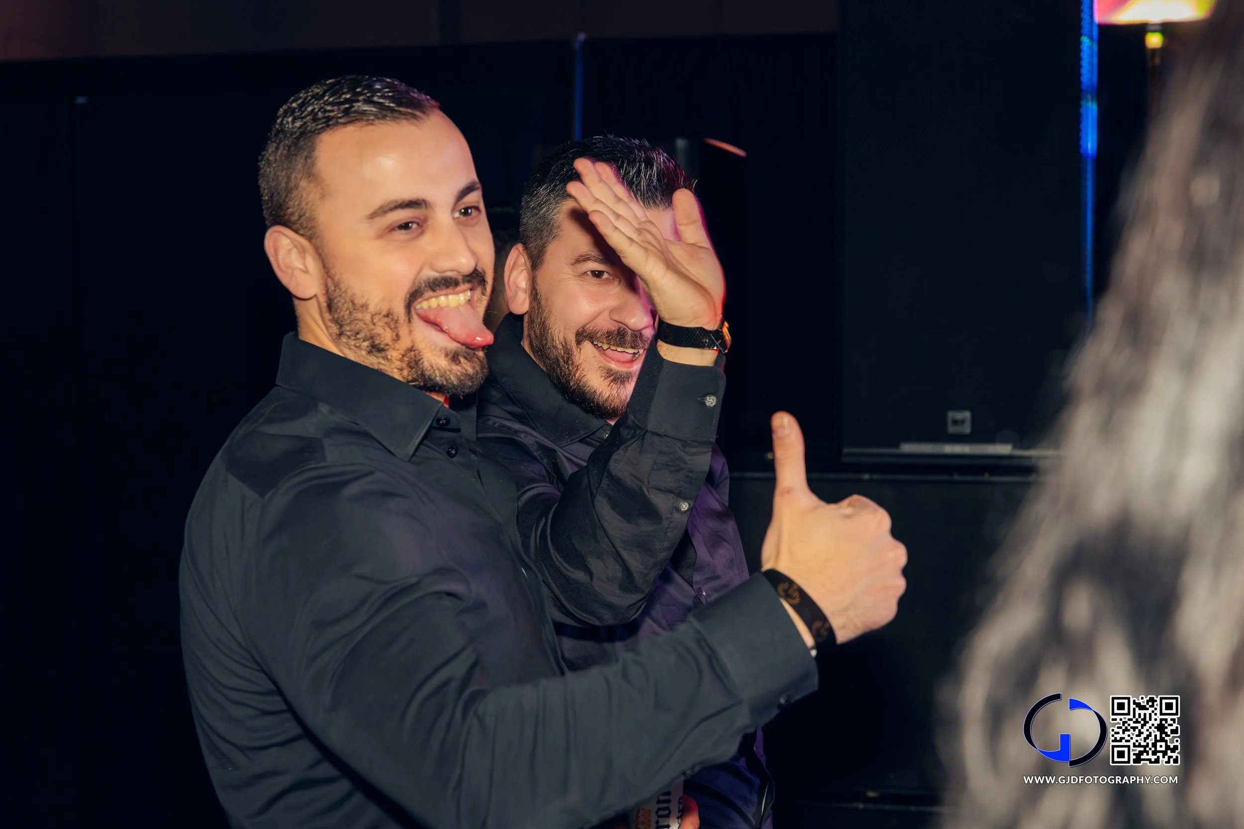 Two men celebrating at a social event, one sticking out his tongue and the other giving a thumbs-up, in a dark room with black background.