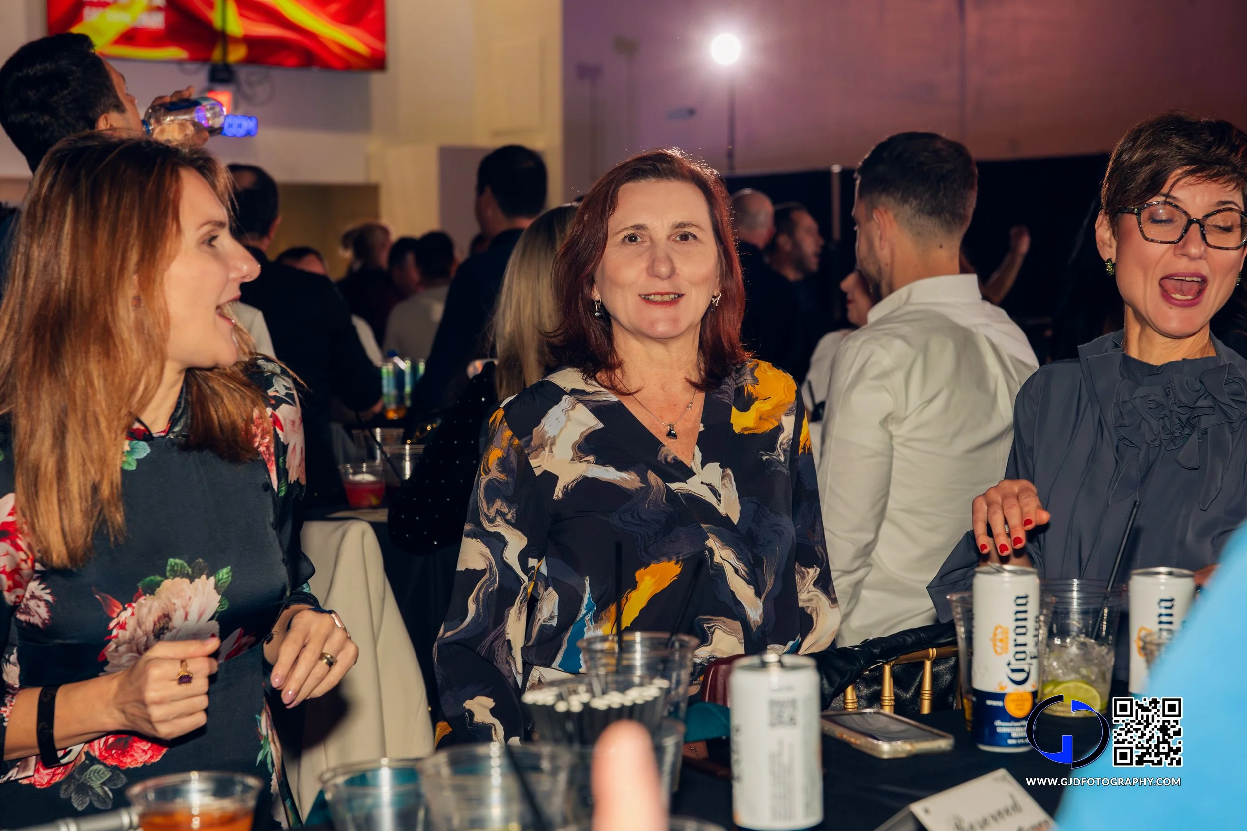 Women socializing at a lively party or gathering, sitting at a table with drinks and a crowded background.