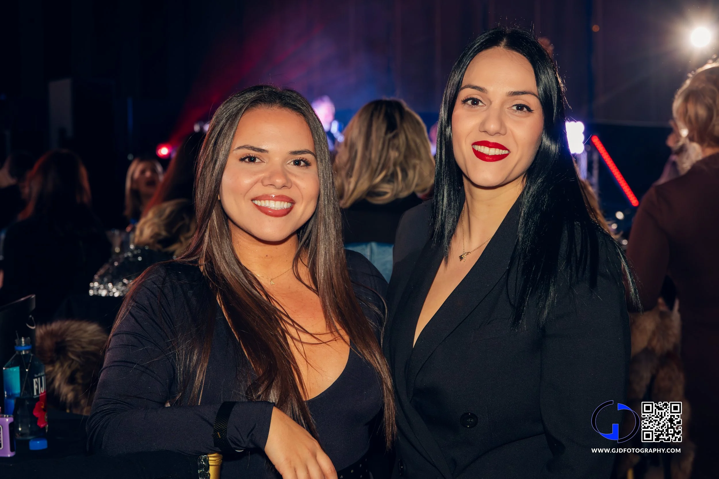 Two women smiling at a nighttime event or party, with a band or performers in the background and colorful stage lights.