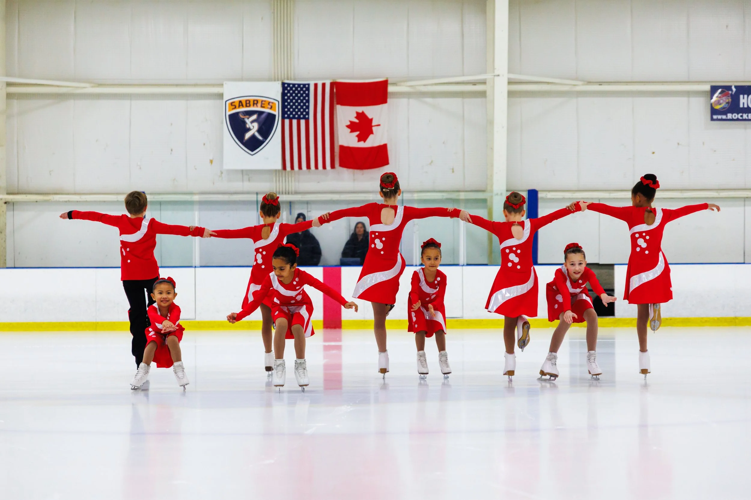 Children in red and white figure skating costumes performing a synchronized routine on an indoor ice rink.