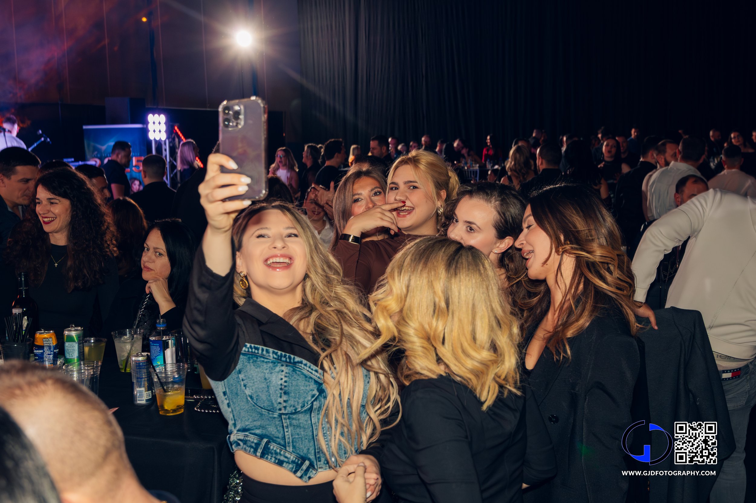 Group of smiling women taking a selfie at a crowded party or event with a stage and lights in background.
