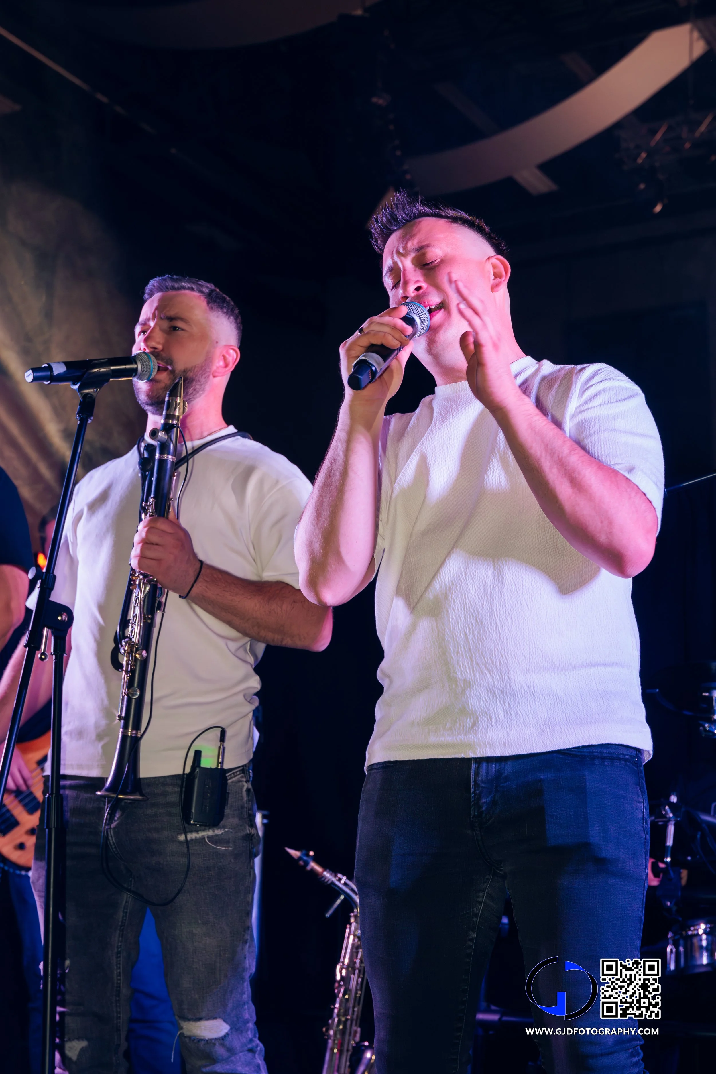 Two male singers performing on stage with one playing a clarinet, under stage lighting. One singer is singing into a microphone with eyes closed, the other is singing into a microphone while holding a clarinet, wearing matching white t-shirts and jea