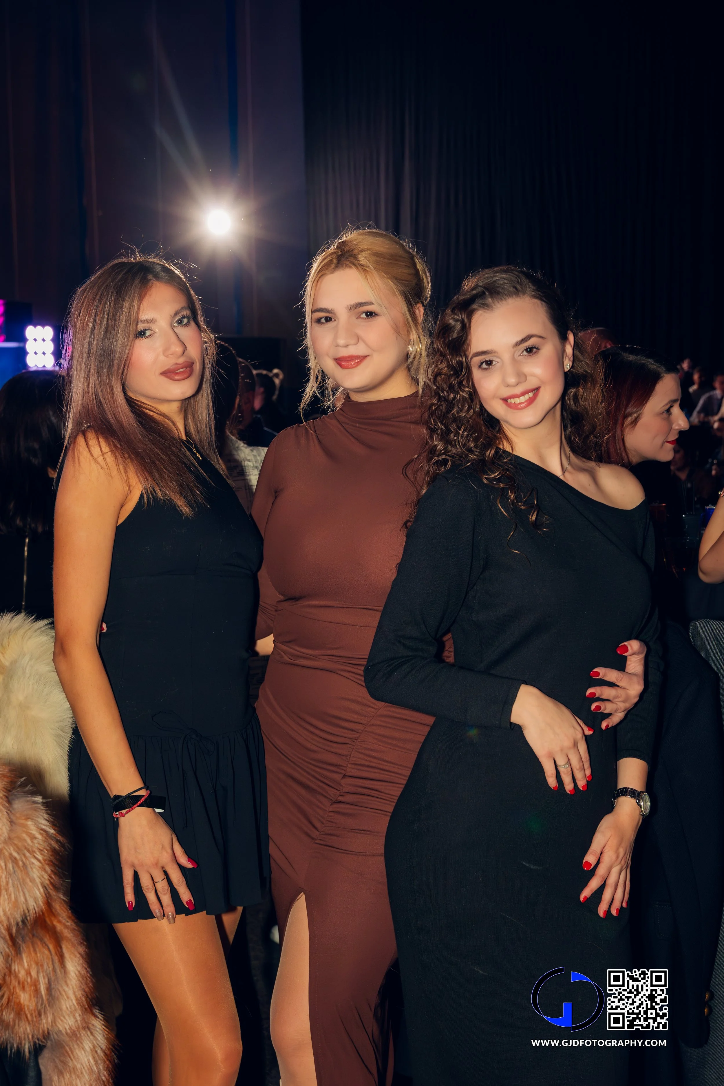 Three women at an indoor event, standing close together and smiling at the camera, with a crowd and stage lights in the background.