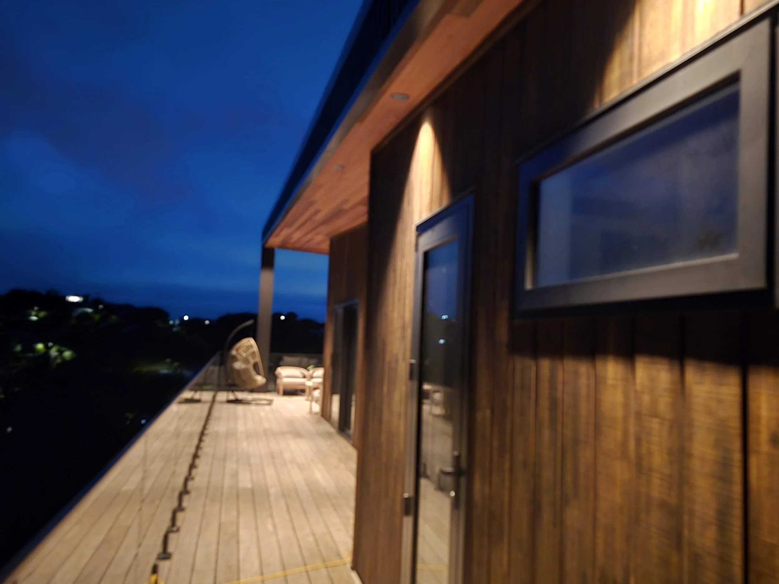 Night view of a wooden deck attached to a modern house with seating and a hanging chair, illuminated by outdoor lights and surrounded by a dark landscape.