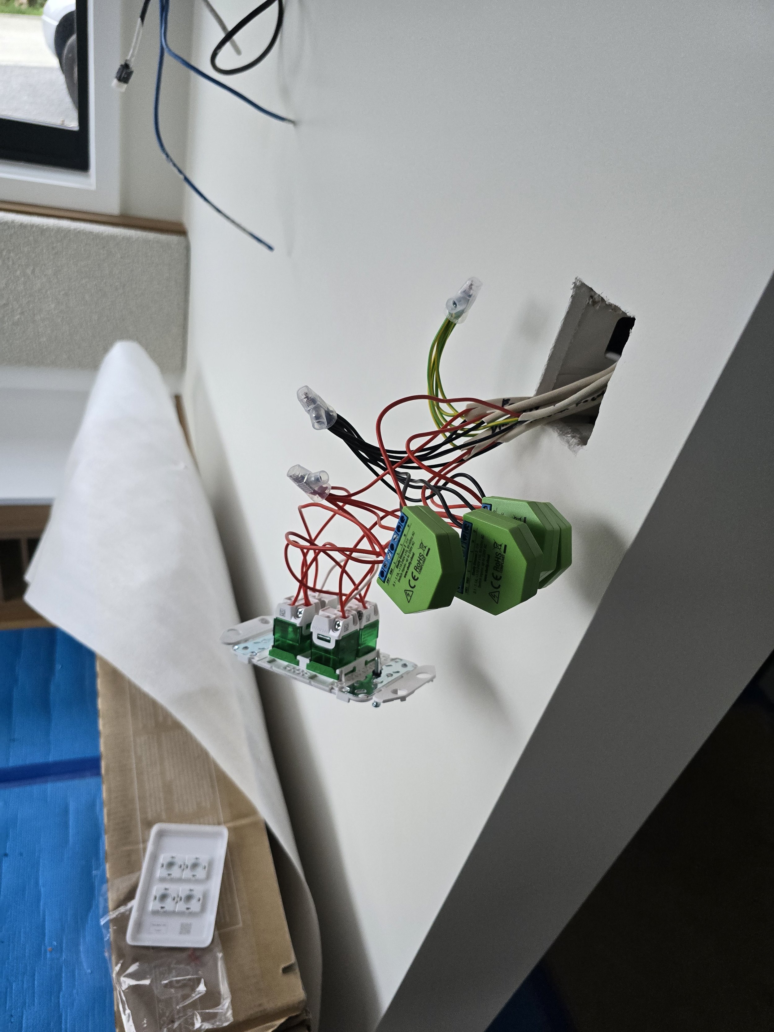 Electrical wiring exposed from a wall with connectors and green components, next to a light switch panel on a cardboard box.