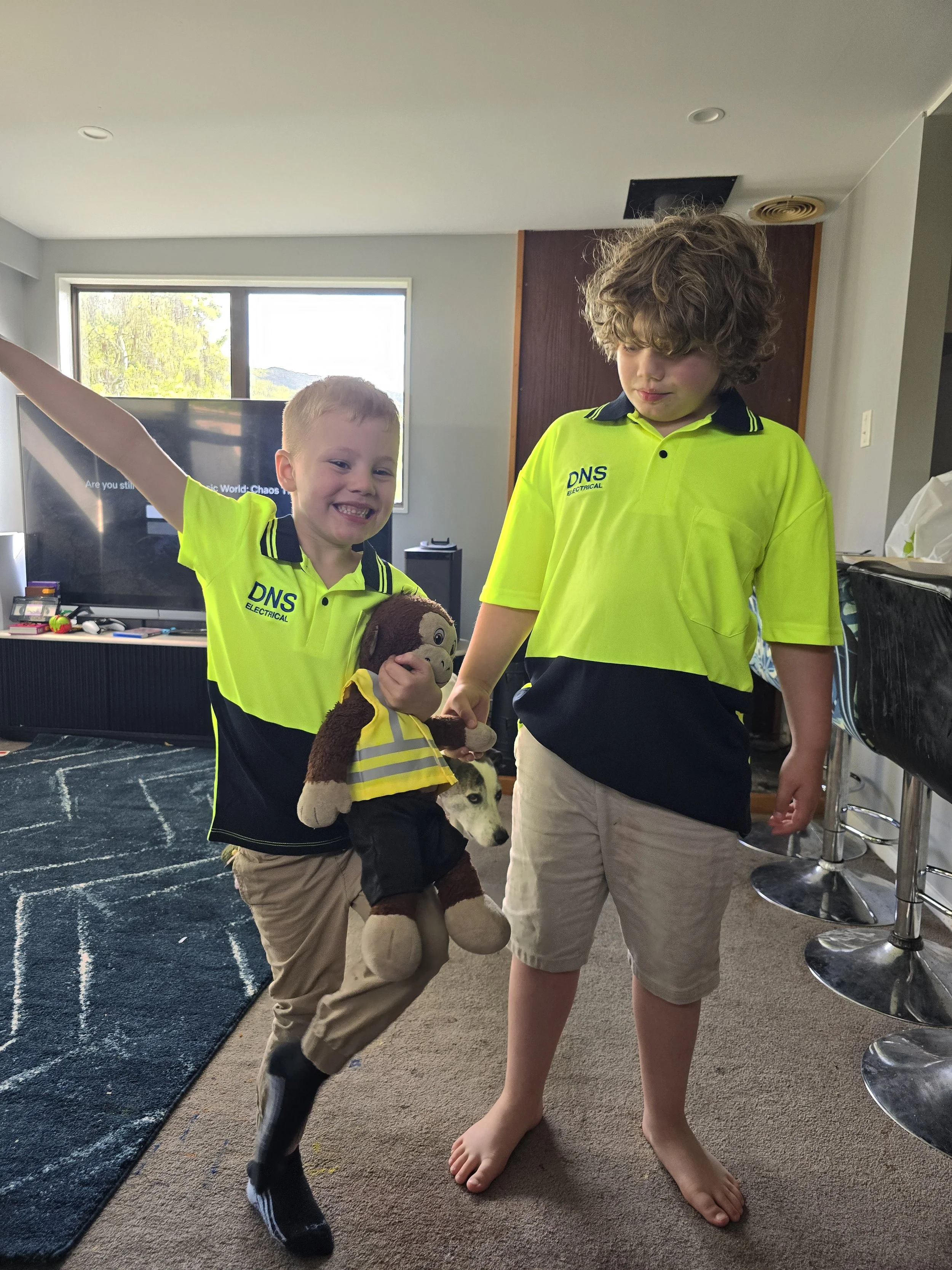 Two children wearing yellow and black shirts with "DNS Electrical" logos, smiling indoors. One child is holding a plush toy wearing a reflective safety vest. A dog is partially visible in the background.