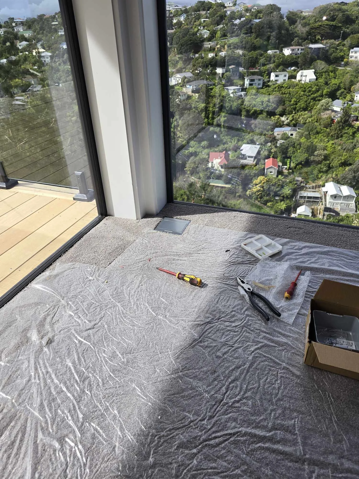 A room under renovation with a large window overlooking a green, hilly residential area. Tools such as screwdrivers and pliers are on a protective sheet covering the floor. A small cardboard box is also visible.