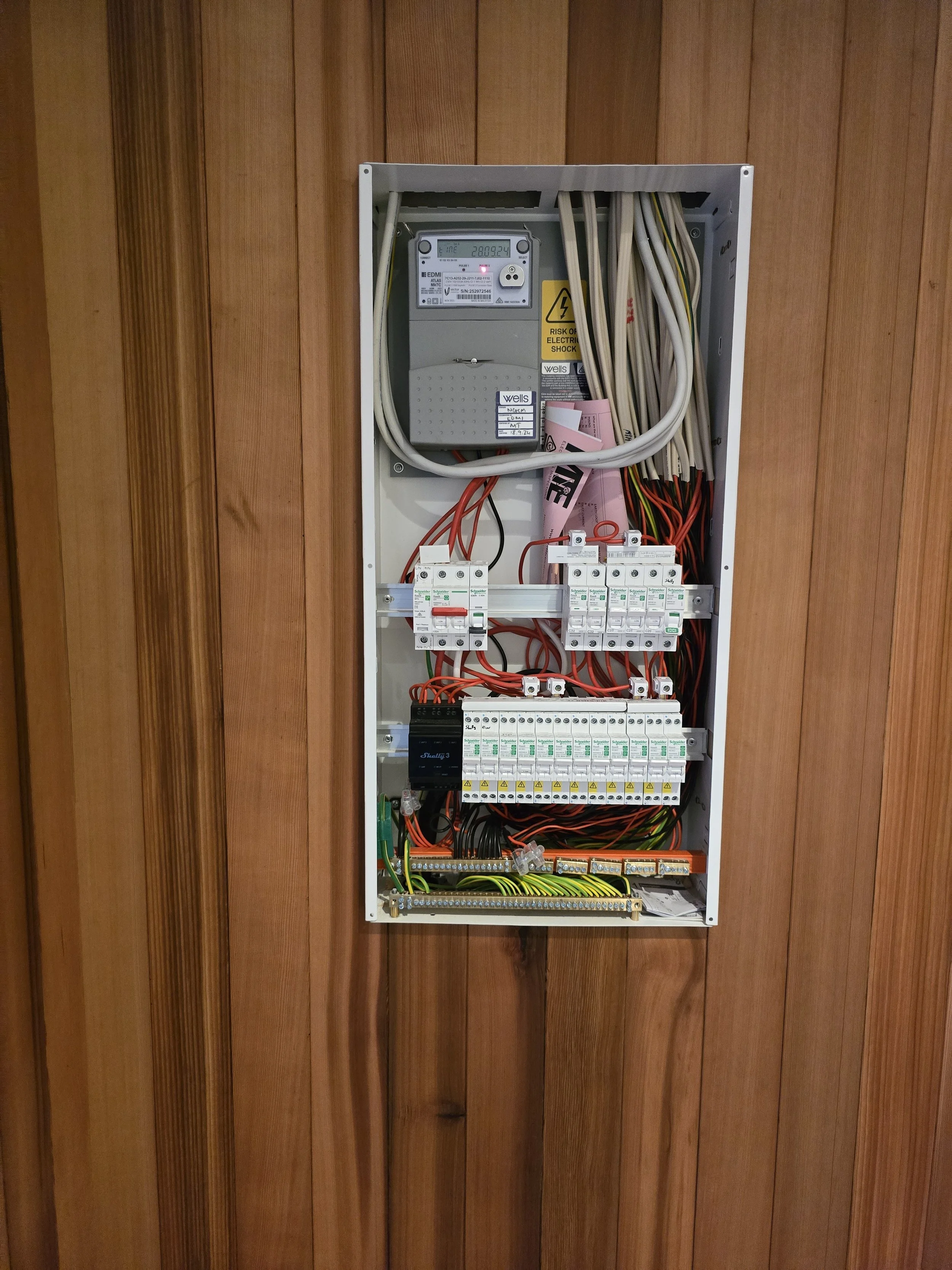 Open electrical panel with wires and circuit breakers on a wooden wall.