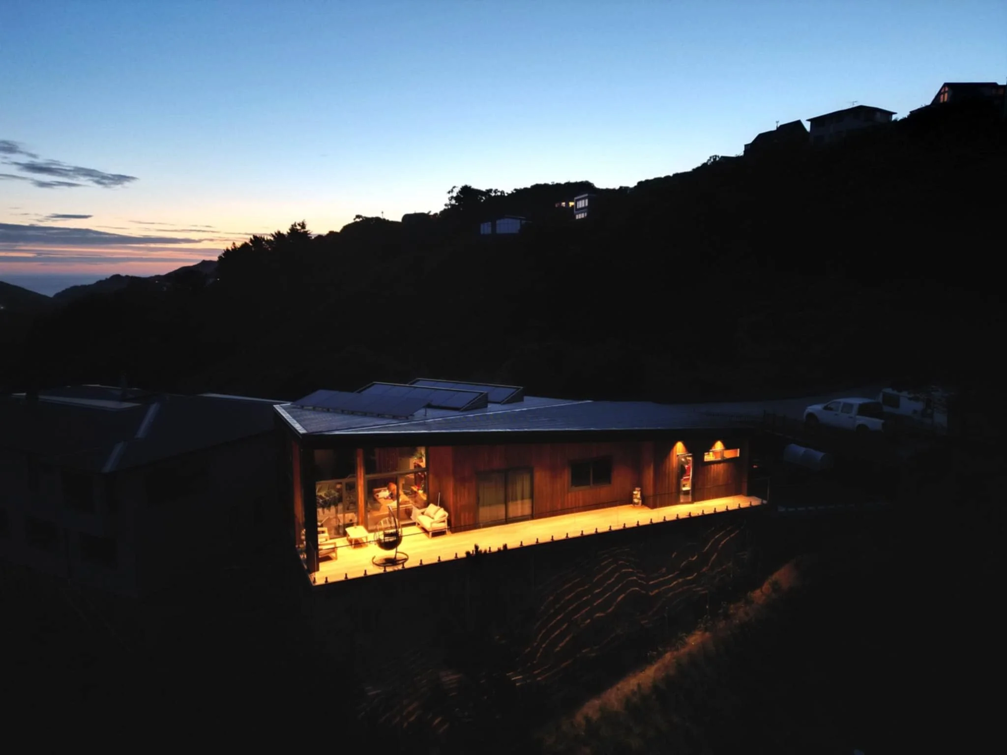 A well-lit modern house on a hill at dusk, surrounded by hills and silhouetted trees, with a visible patio and interior lights glowing.