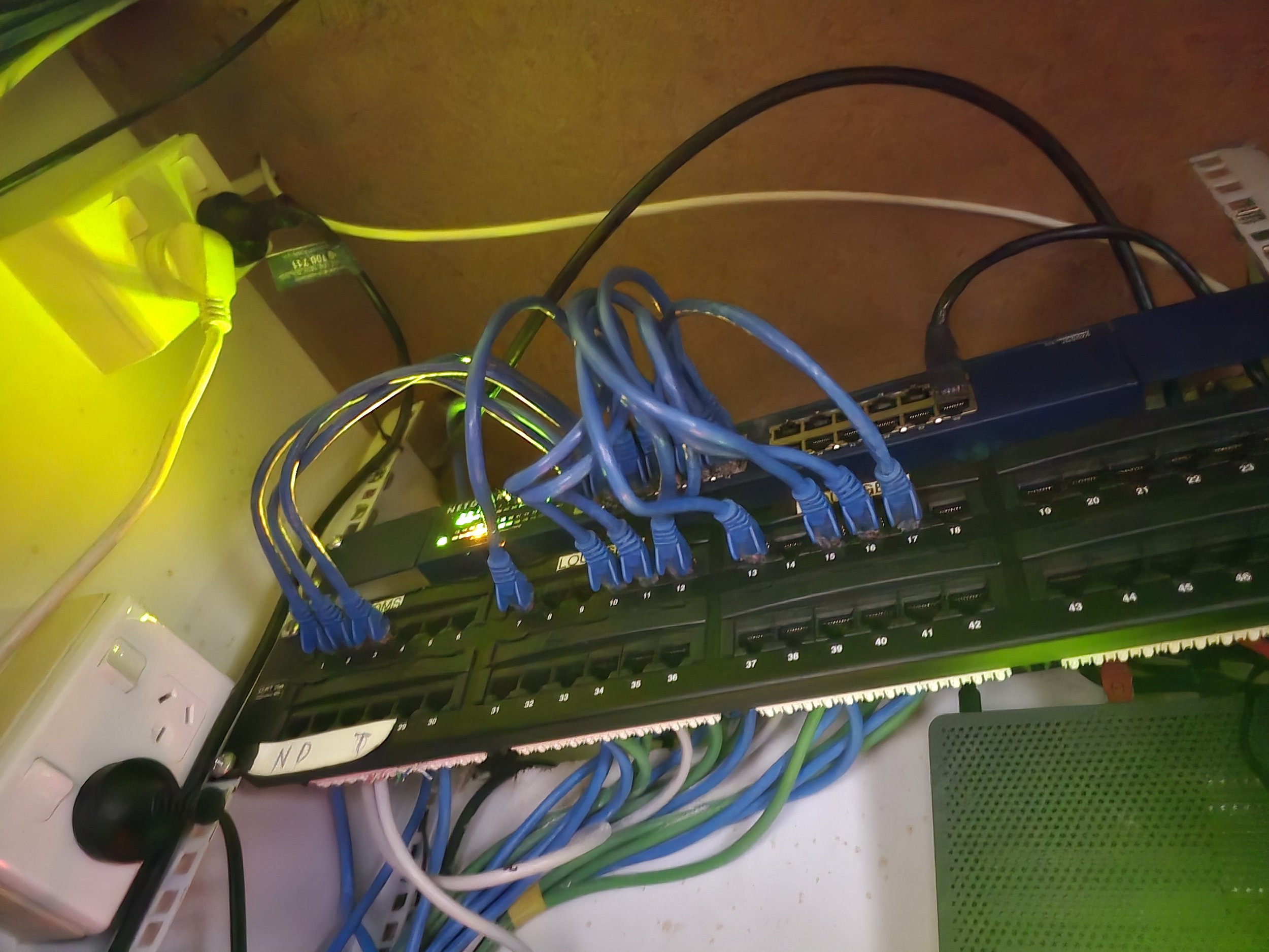 Network patch panel with blue Ethernet cables connected, alongside a network switch with green indicator lights. Electrical outlet and plugs are visible in the background.