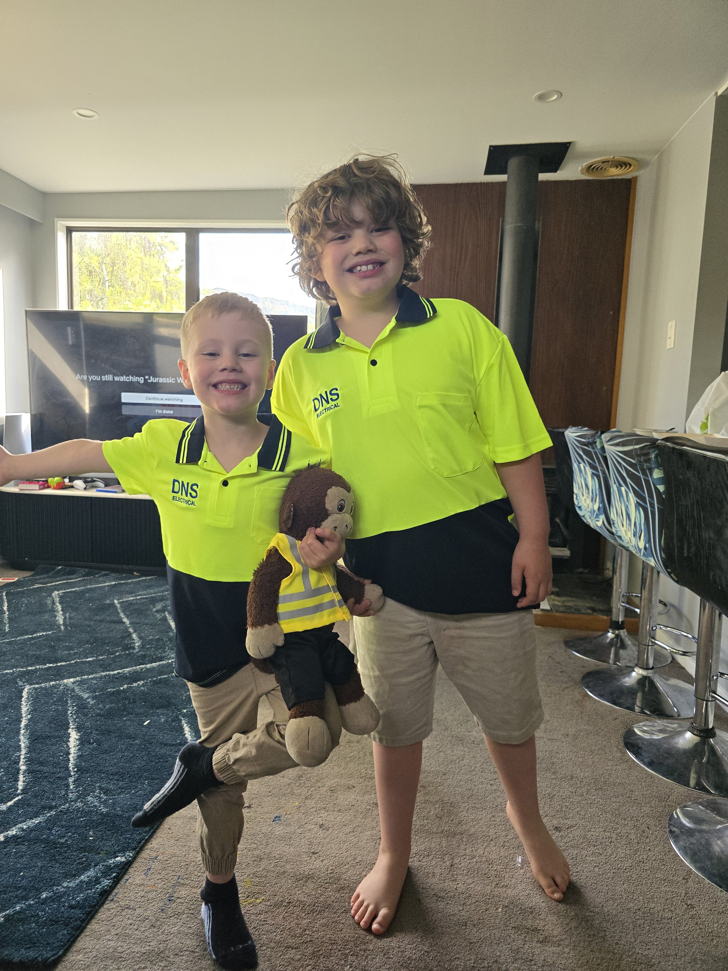 Two children wearing yellow shirts smiling indoors, one holding a stuffed monkey toy dressed in a matching outfit. A television is in the background.