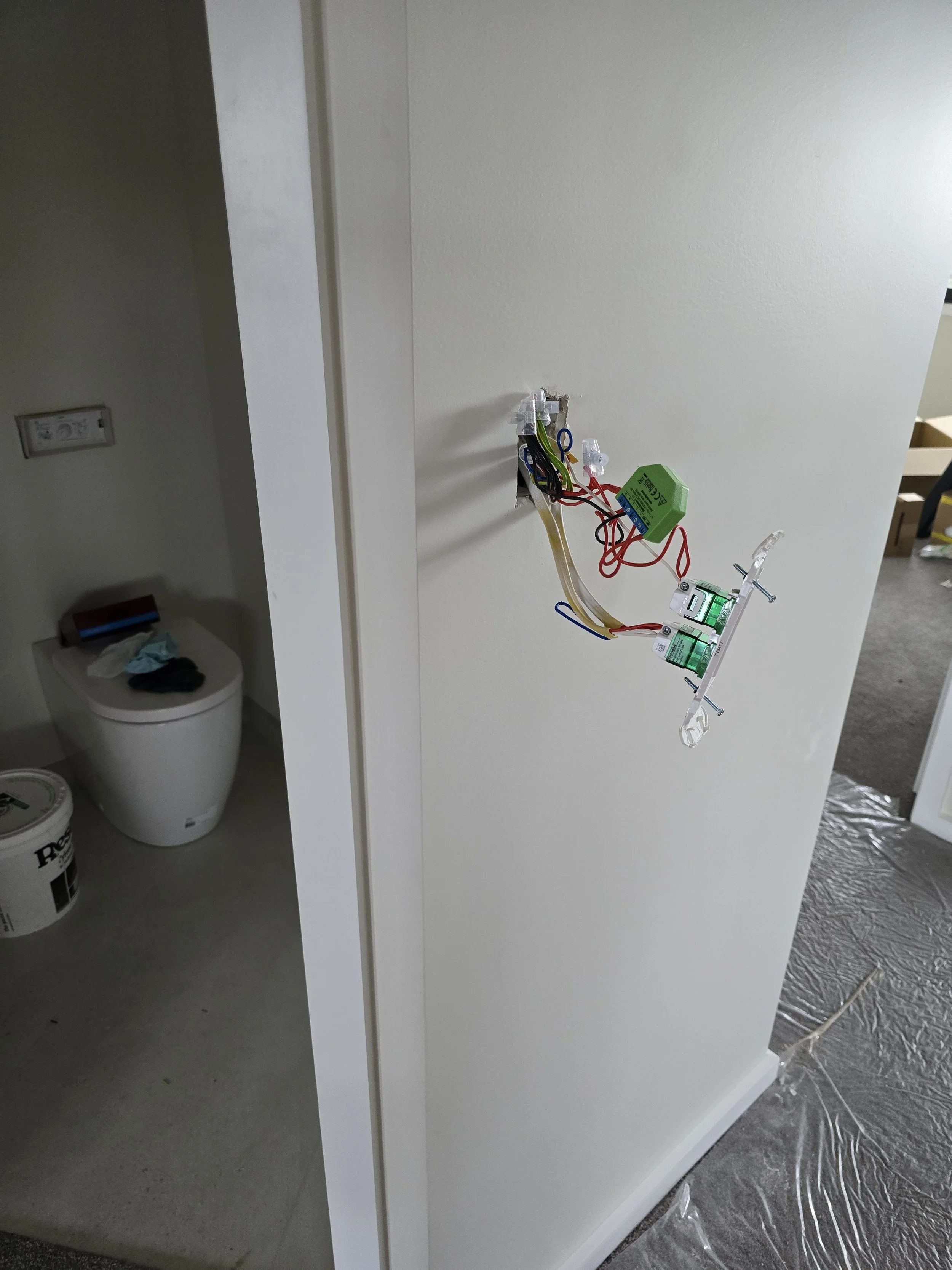 A partially installed light switch with exposed wiring on a wall. Nearby, a bucket of paint and cleaning supplies are on the floor, indicating ongoing renovations. A toilet is visible in an adjacent room.