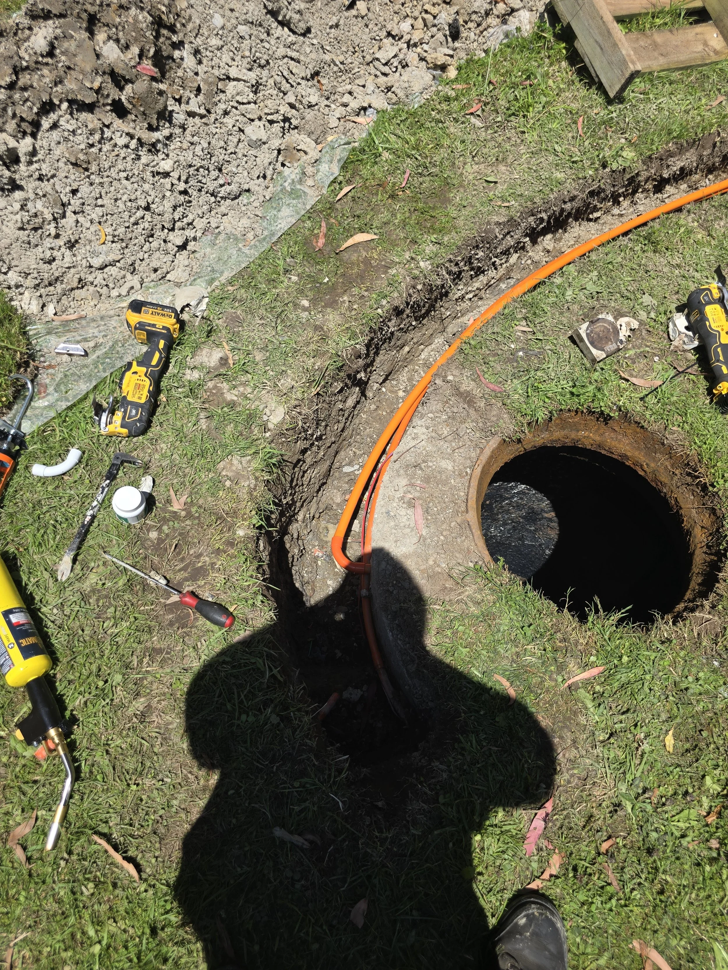 A construction site with a circular manhole, an orange conduit, tools, and a worker's shadow on grass.
