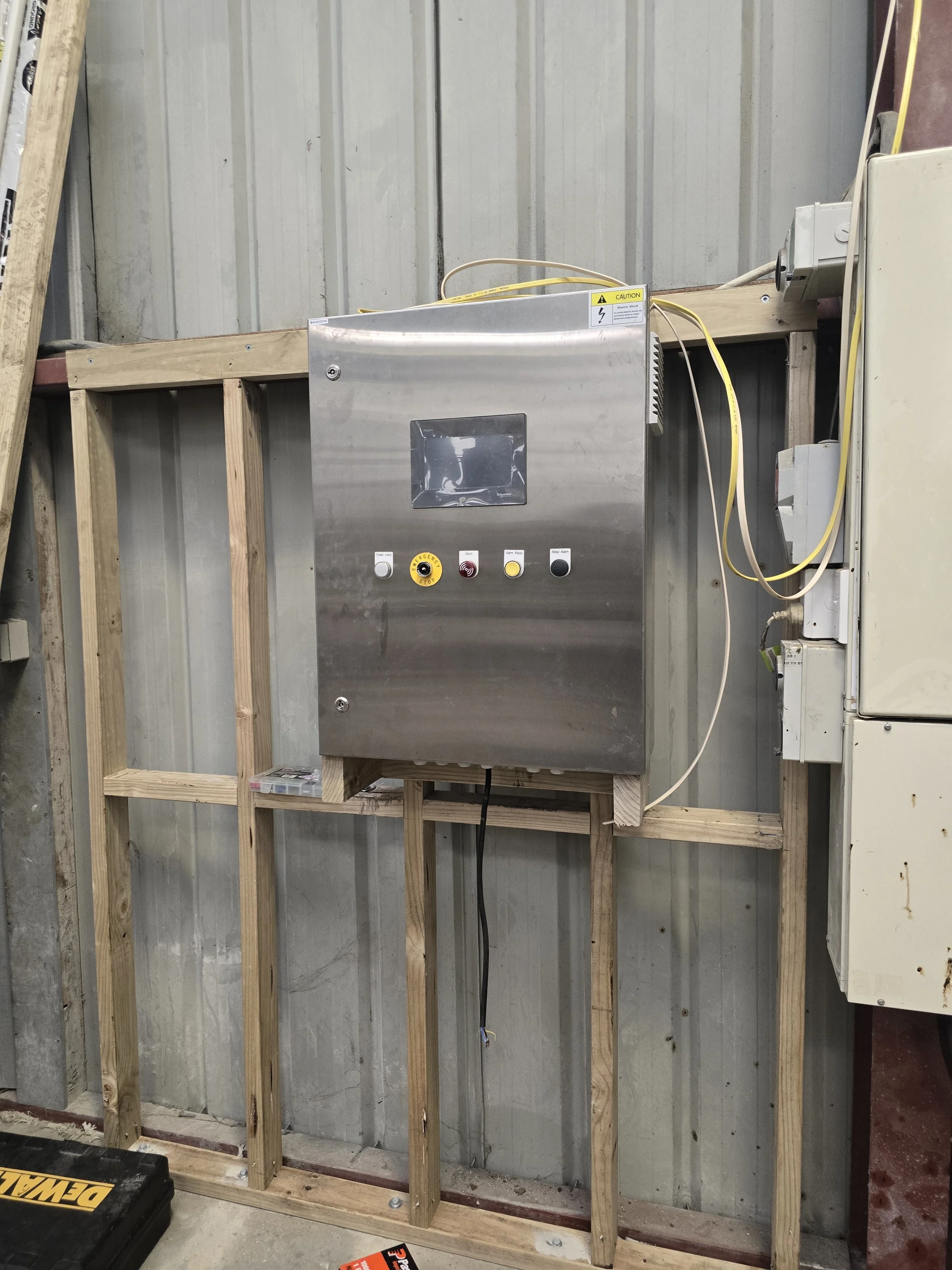 Metal electrical control panel mounted on a wooden frame with buttons and wires, inside a warehouse or industrial setting.