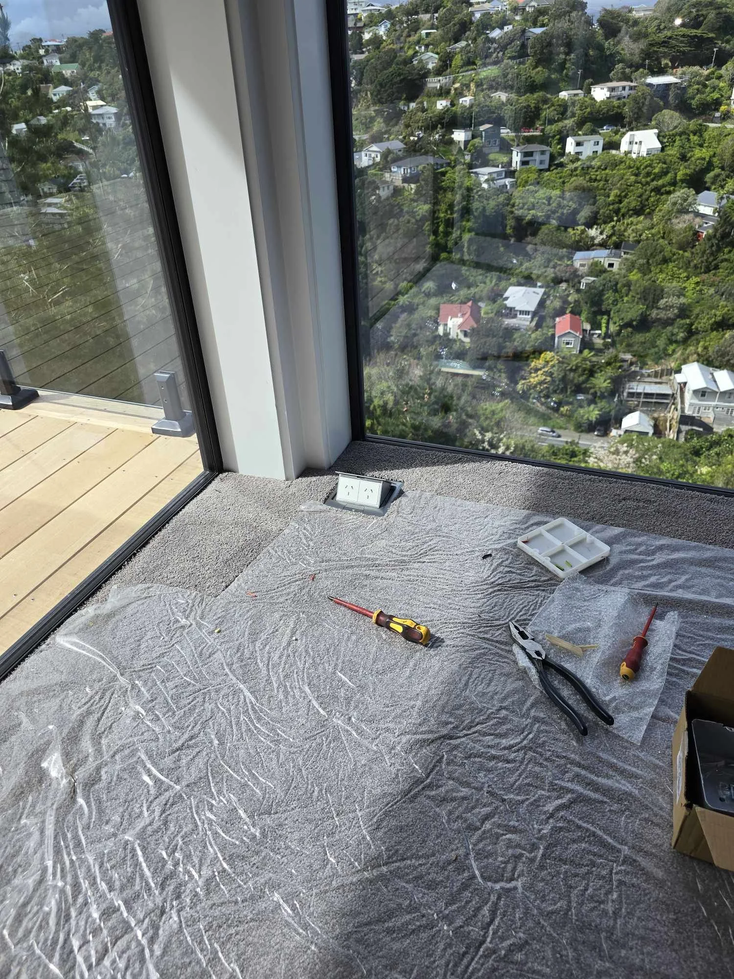 Room corner with large glass windows, covered floor with plastic sheet, tools like screwdriver and pliers, view of green landscape and houses outside.