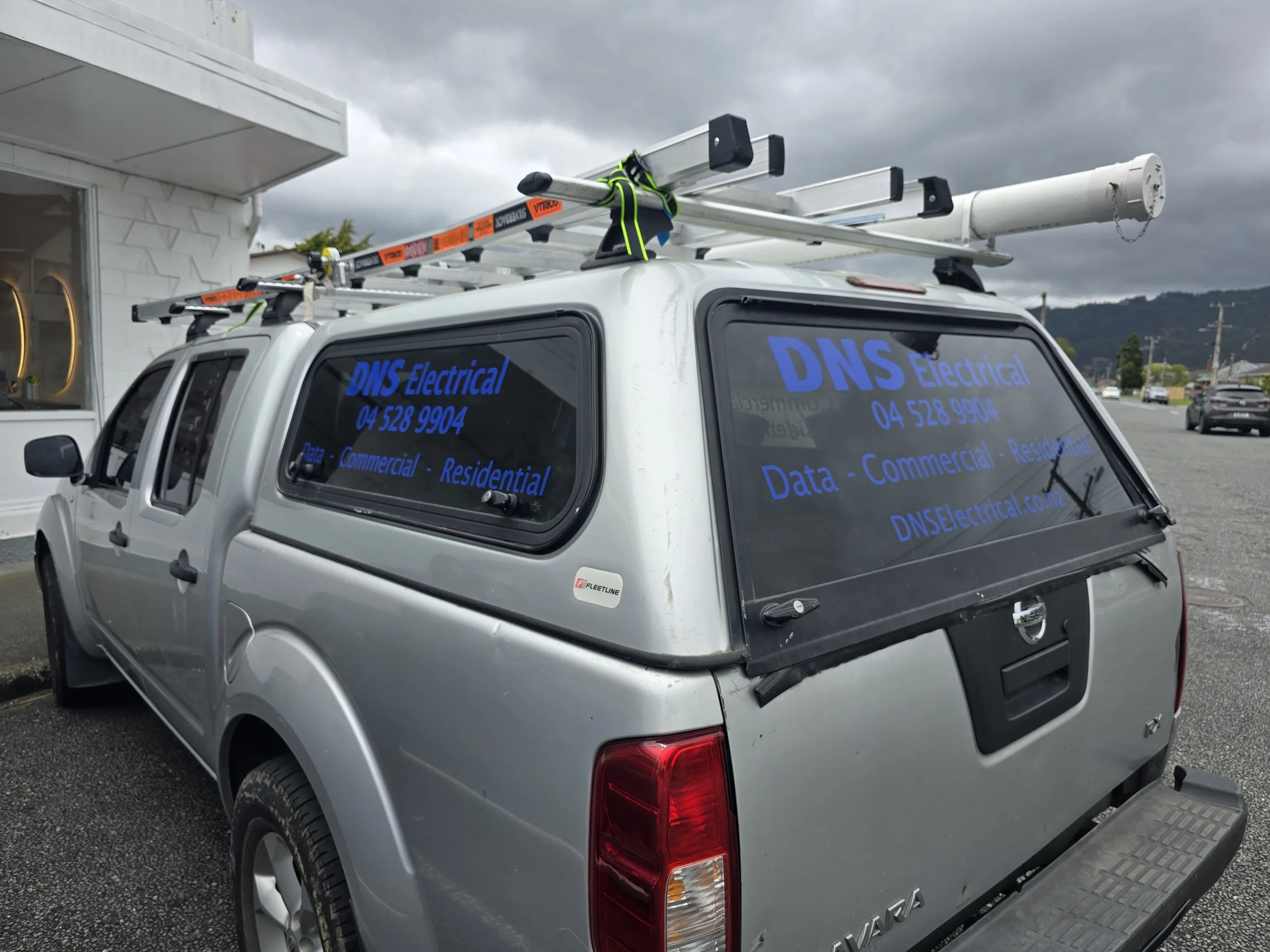 Silver pickup truck with company branding, featuring ladders on the roof, parked outside a building.