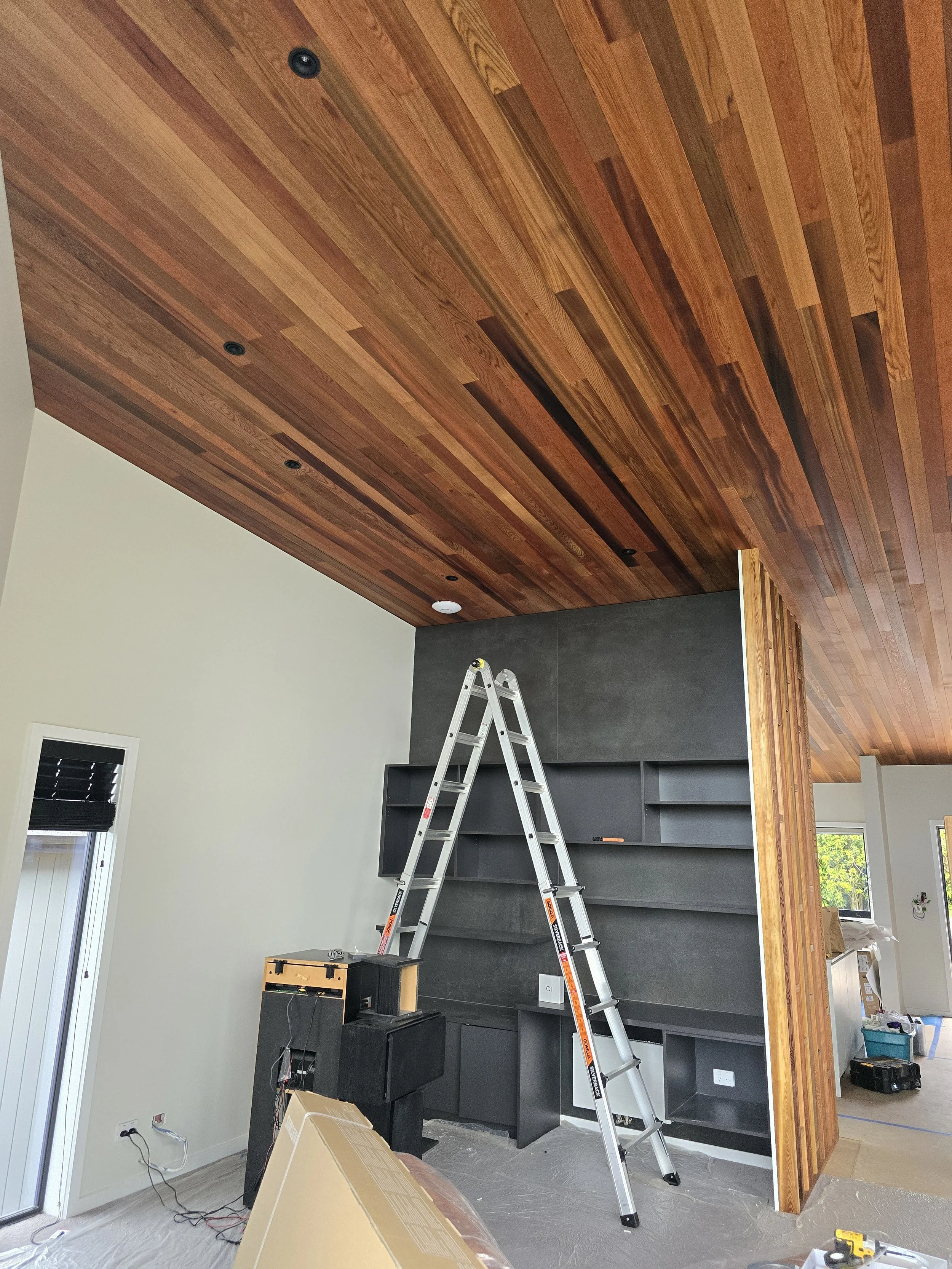 Interior room under construction with a wooden ceiling, a ladder leaning against a dark shelving unit, and a partially assembled area with tools and boxes on the floor.