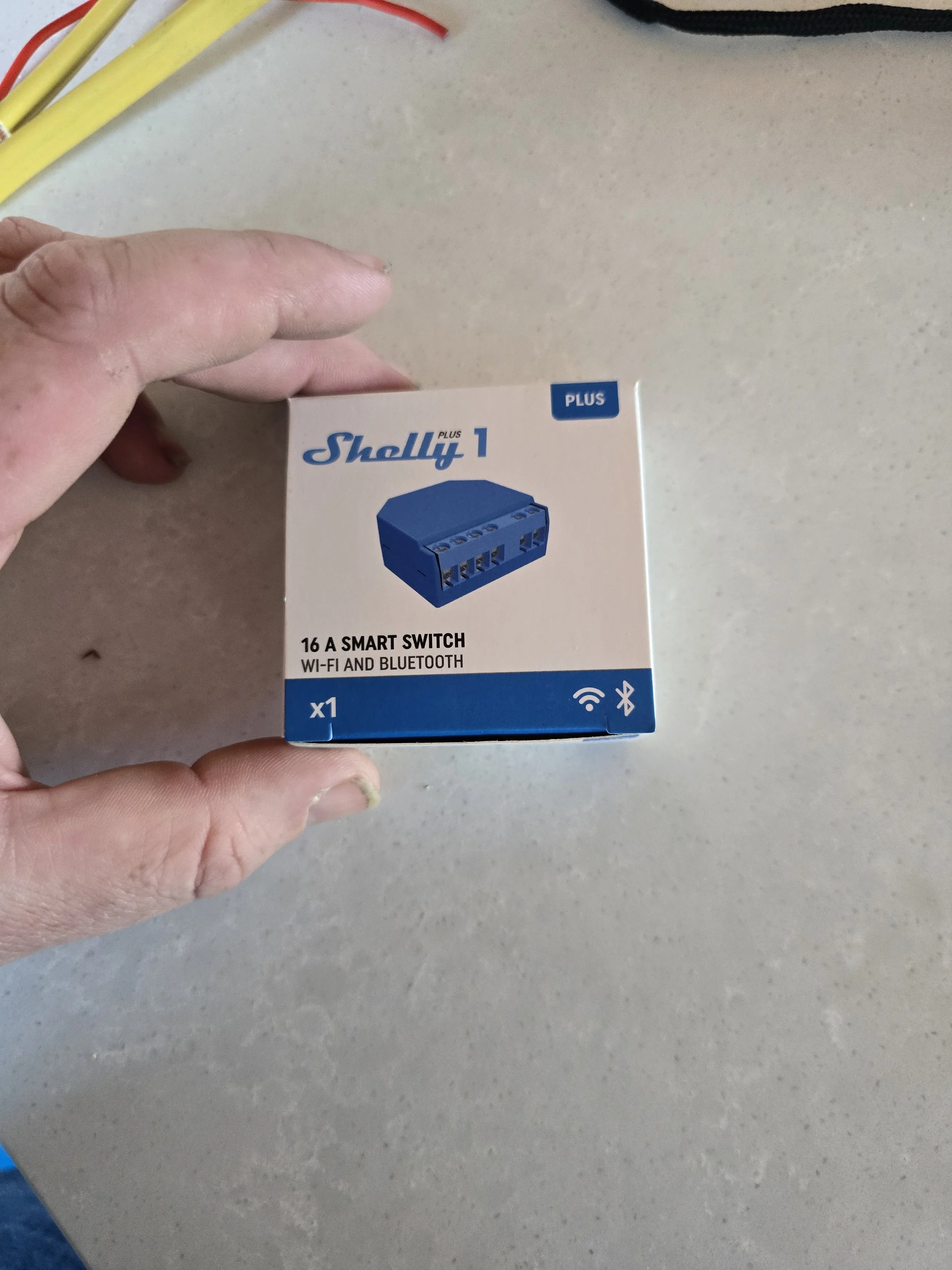 A person holding a Shelly Plus 1 smart switch box on a table. The box indicates the device is a 16A smart switch with Wi-Fi and Bluetooth capabilities.