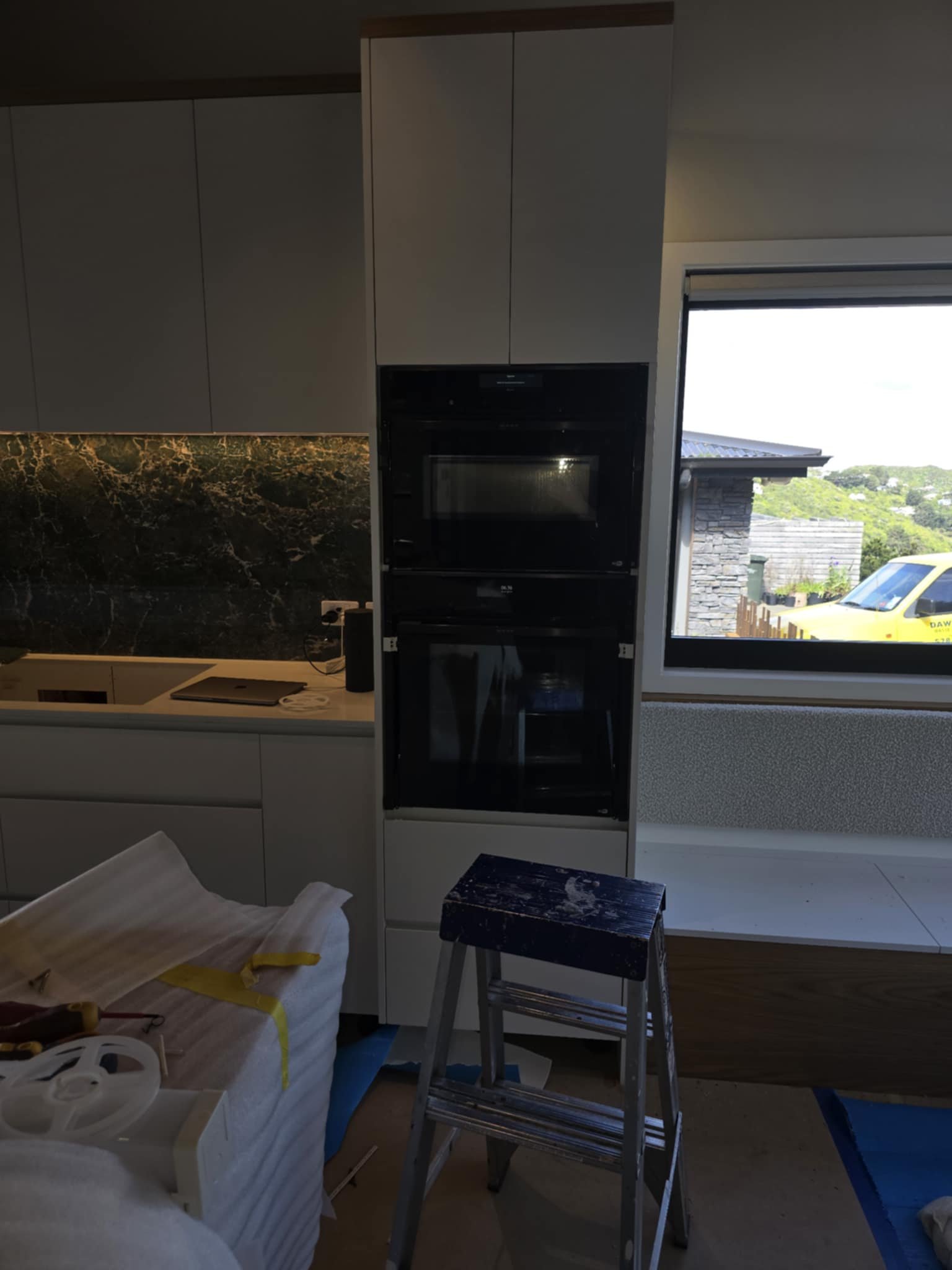 Modern kitchen under construction with a built-in oven, white cabinets, a dark stone backsplash, a countertop with a sink and stovetop, and a window. There is a step ladder and unpacked furniture in the foreground.