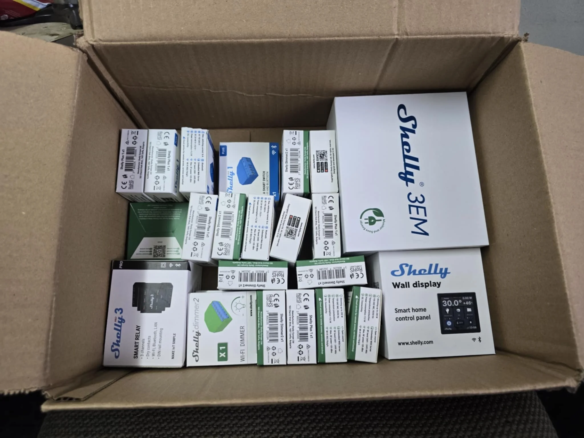 Cardboard box containing multiple Shelly smart home device packages, including Shelly 3EM, Shelly Wall Display, and Shelly Dimmer 2.