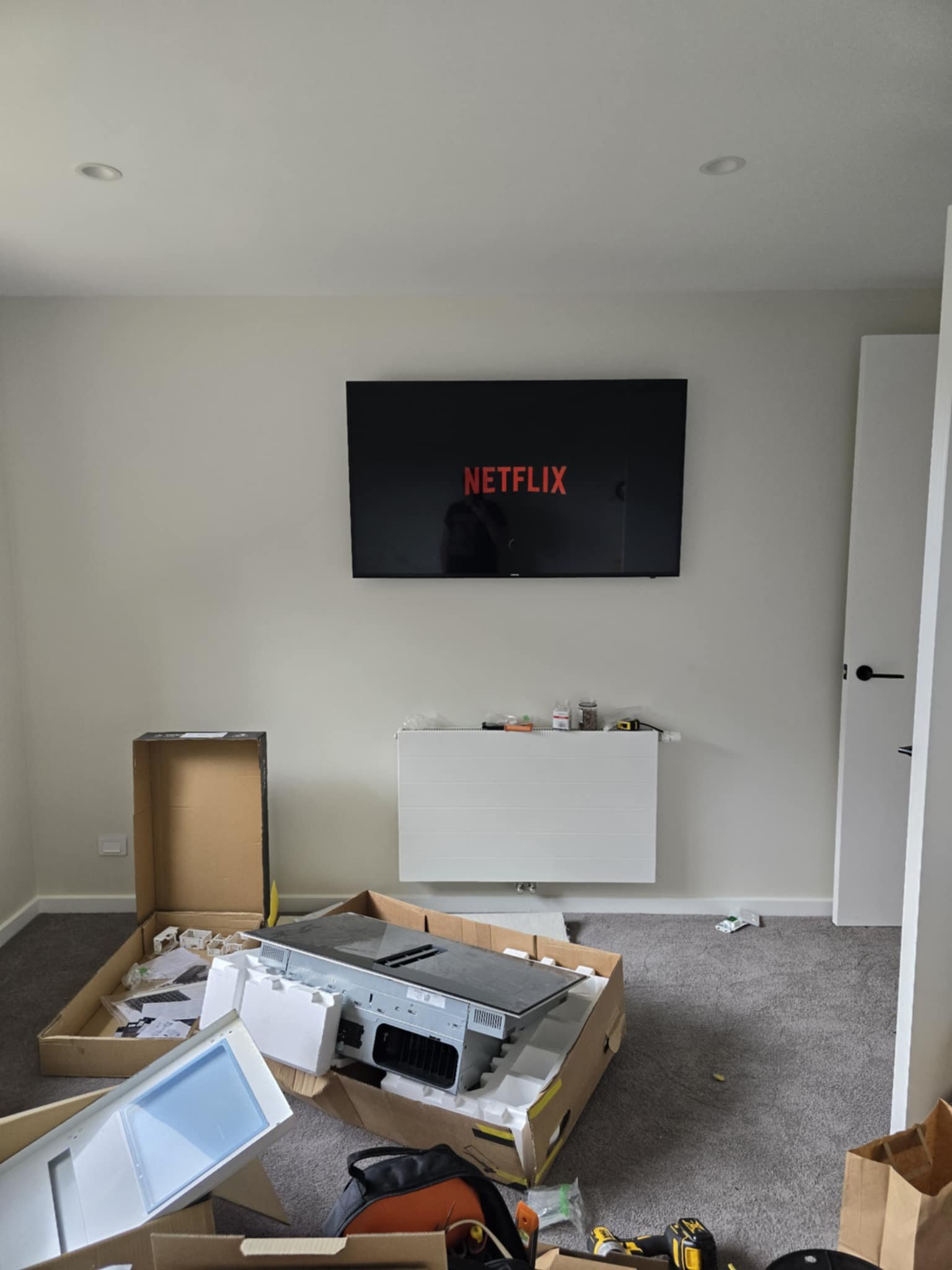 Room with a wall-mounted TV displaying the Netflix logo, surrounded by unpacked cardboard boxes and scattered items, suggesting recent setup or installation.