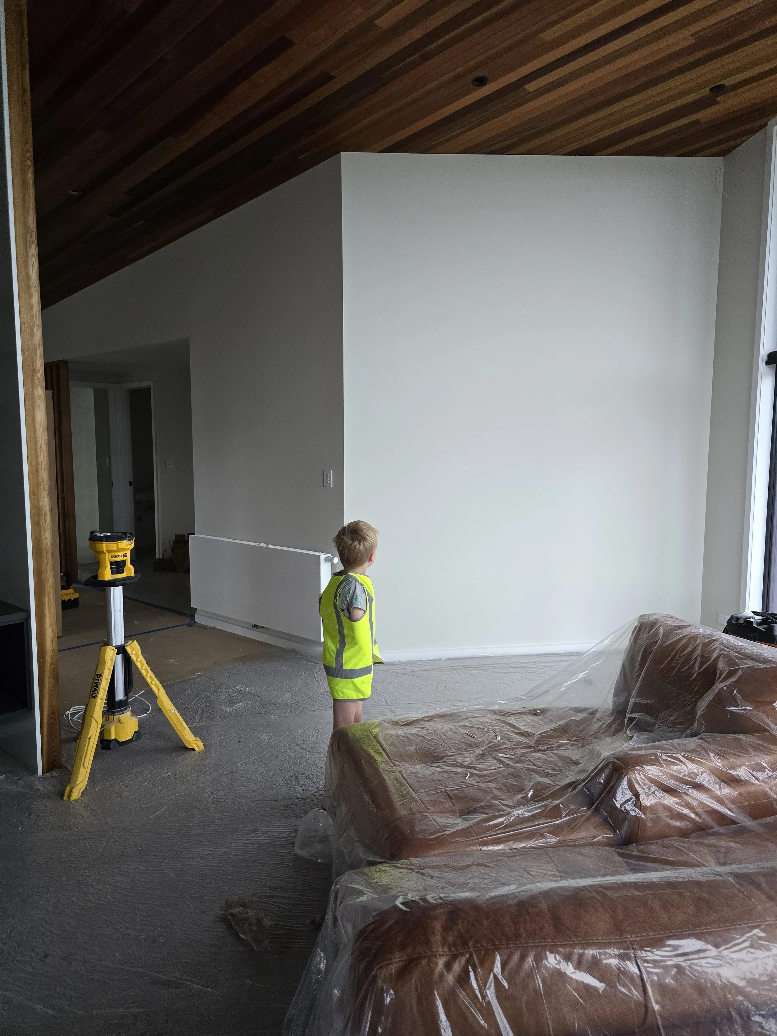 A child in a safety vest stands in a partially renovated room with plastic-wrapped furniture and tools.
