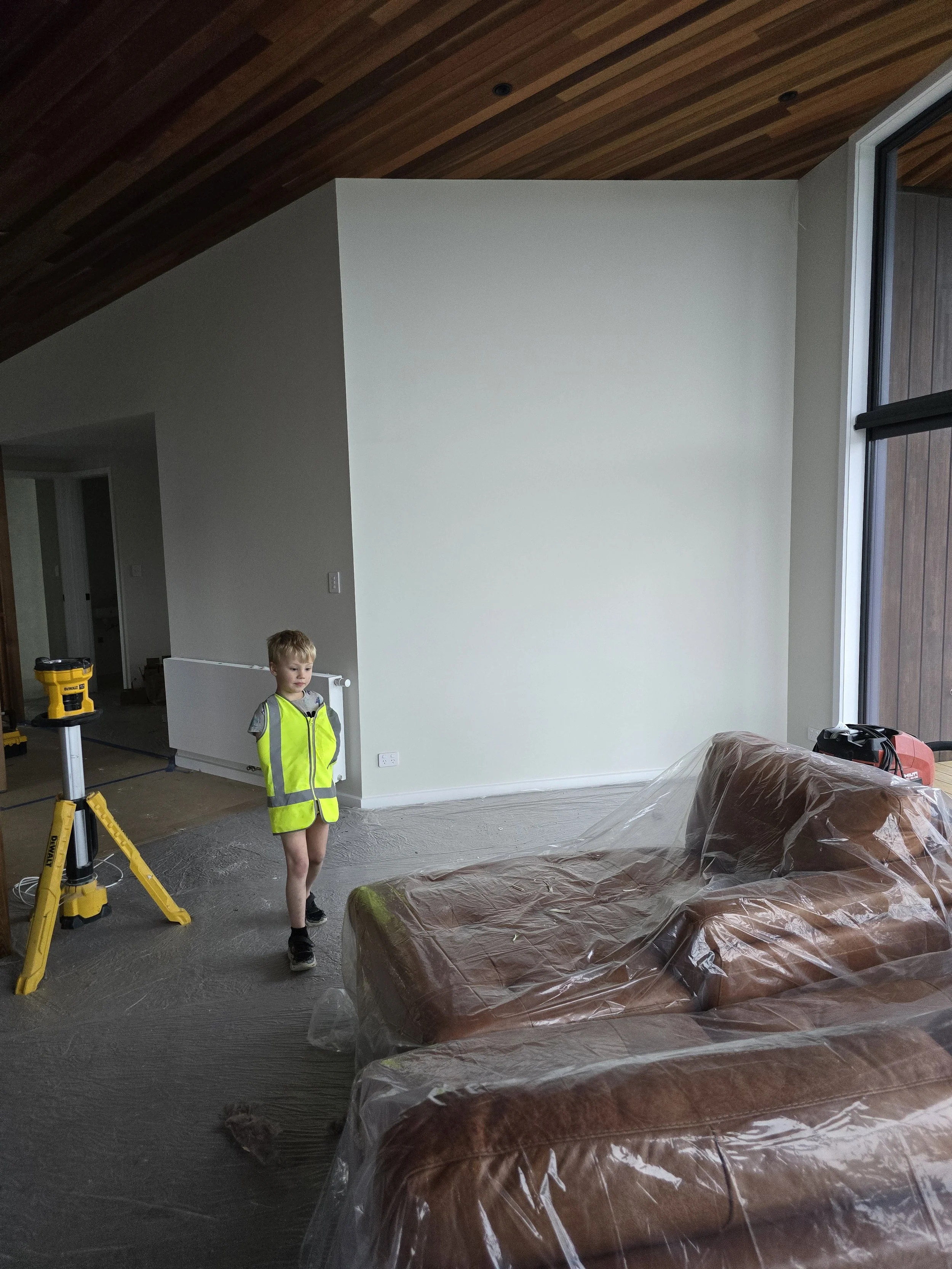 A young child in a bright safety vest stands in a partially furnished room under renovation, with plastic-covered furniture and construction equipment visible.