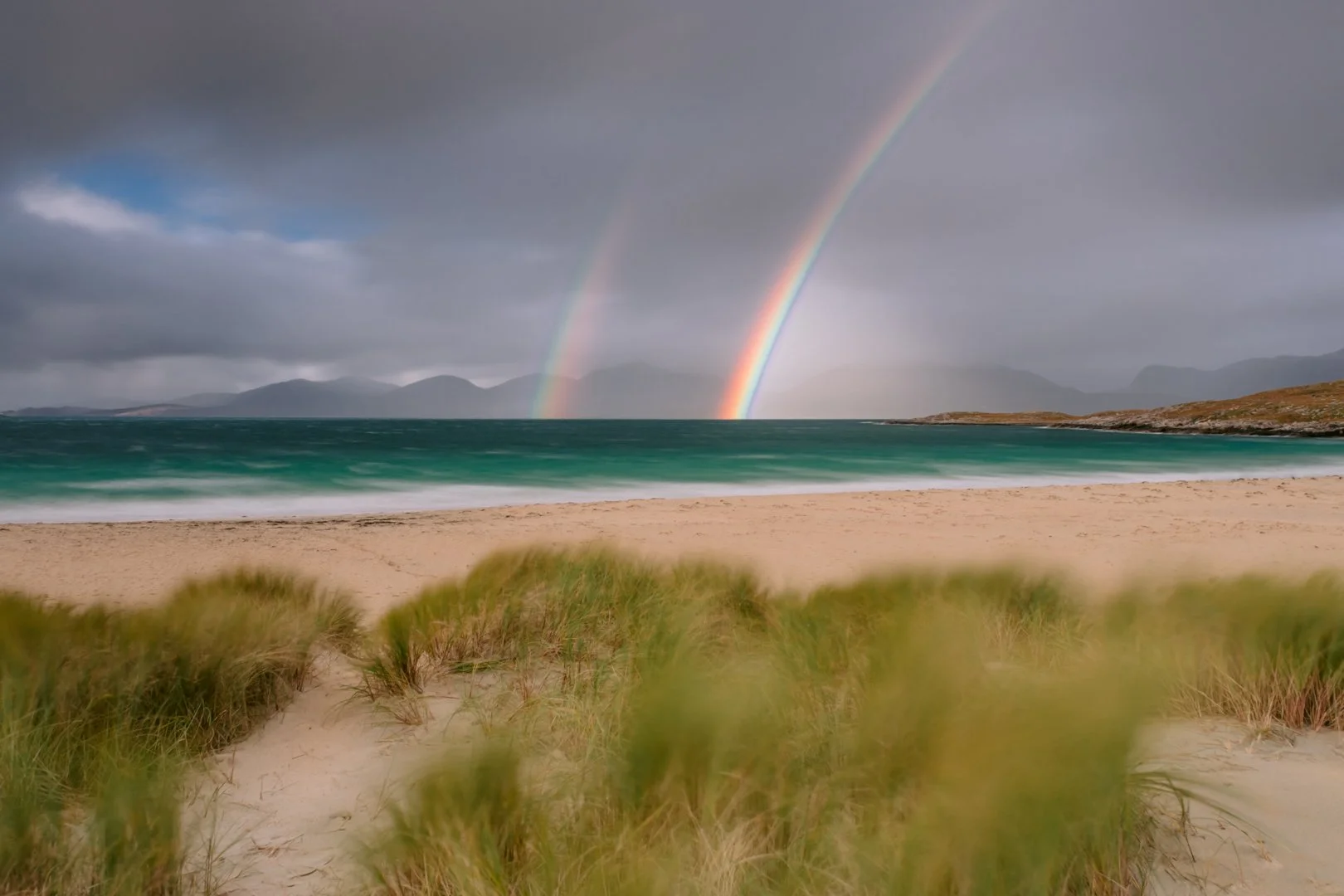 Scenic beach with sand dunes and green grass, dark cloudy sky with a rainbow over the ocean and distant mountains.