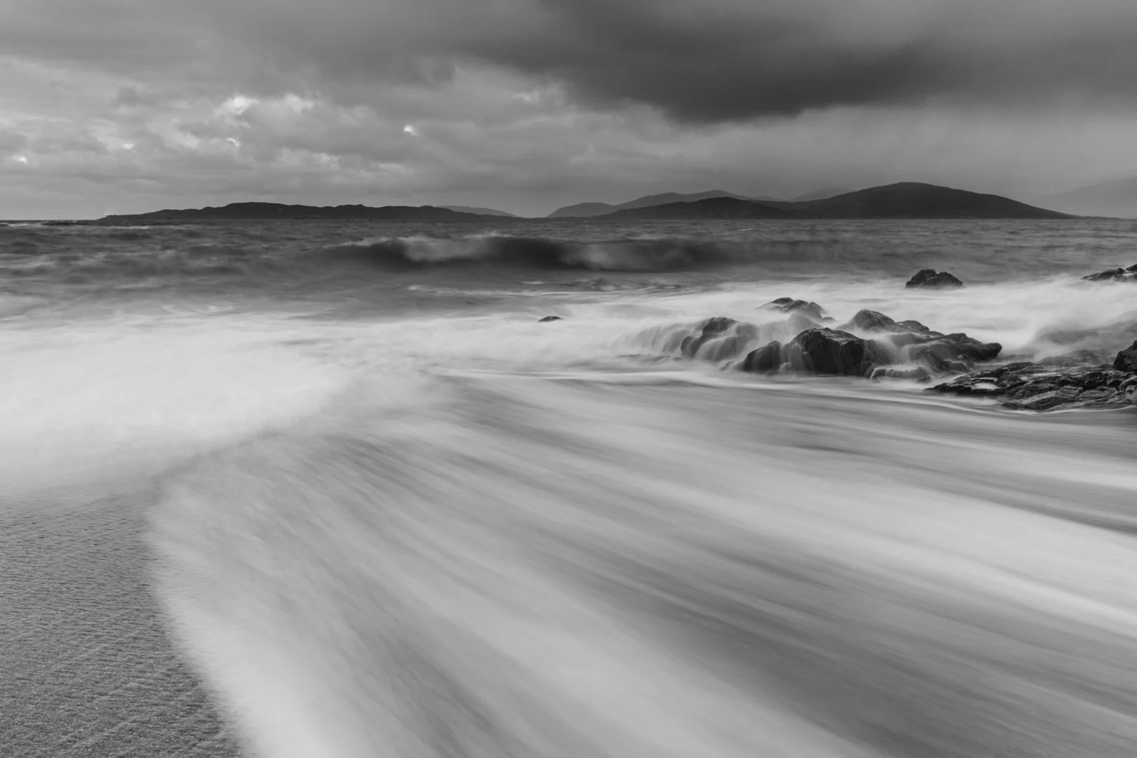 Black and white photo of a beach with waves crashing onto the shore and rocks, with distant islands and cloudy sky in the background.