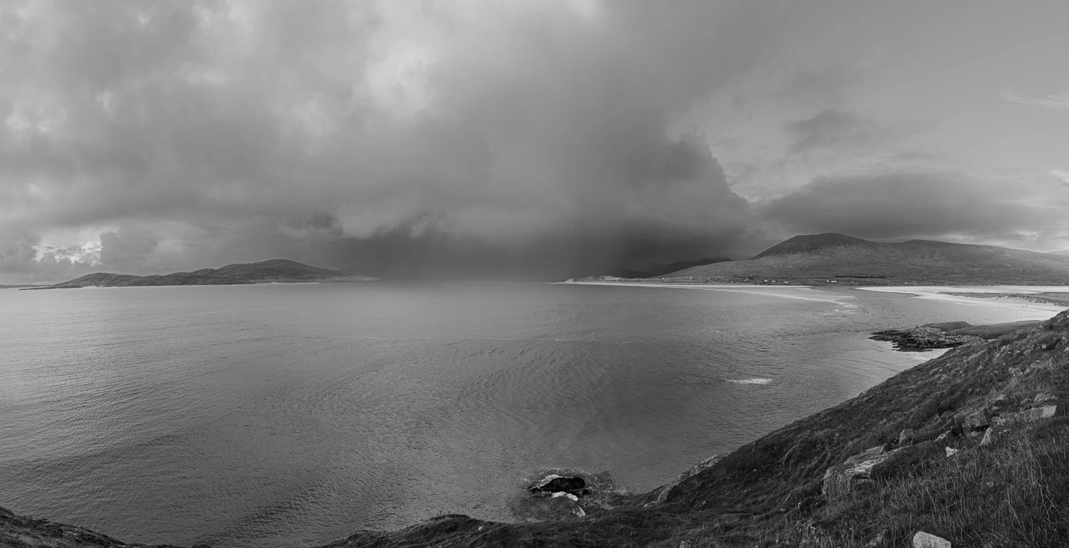 Black and white photo of a beach with water and mountains in the background, under a cloudy sky.